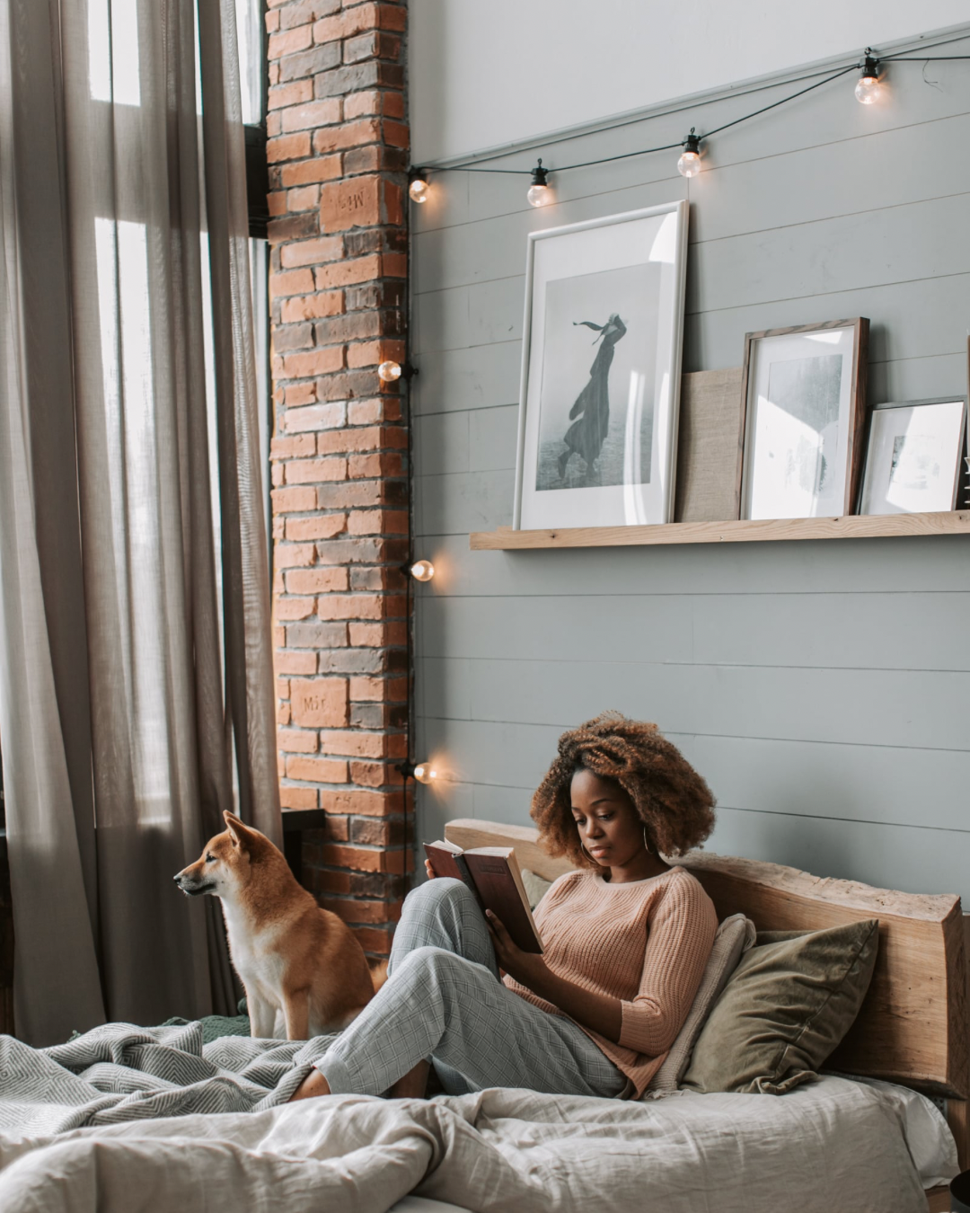 A woman with curly hair reading a book in bed next to a Shiba Inu dog in a cozy bedroom with an exposed brick wall and framed art on a shelf, decorated with string lights.