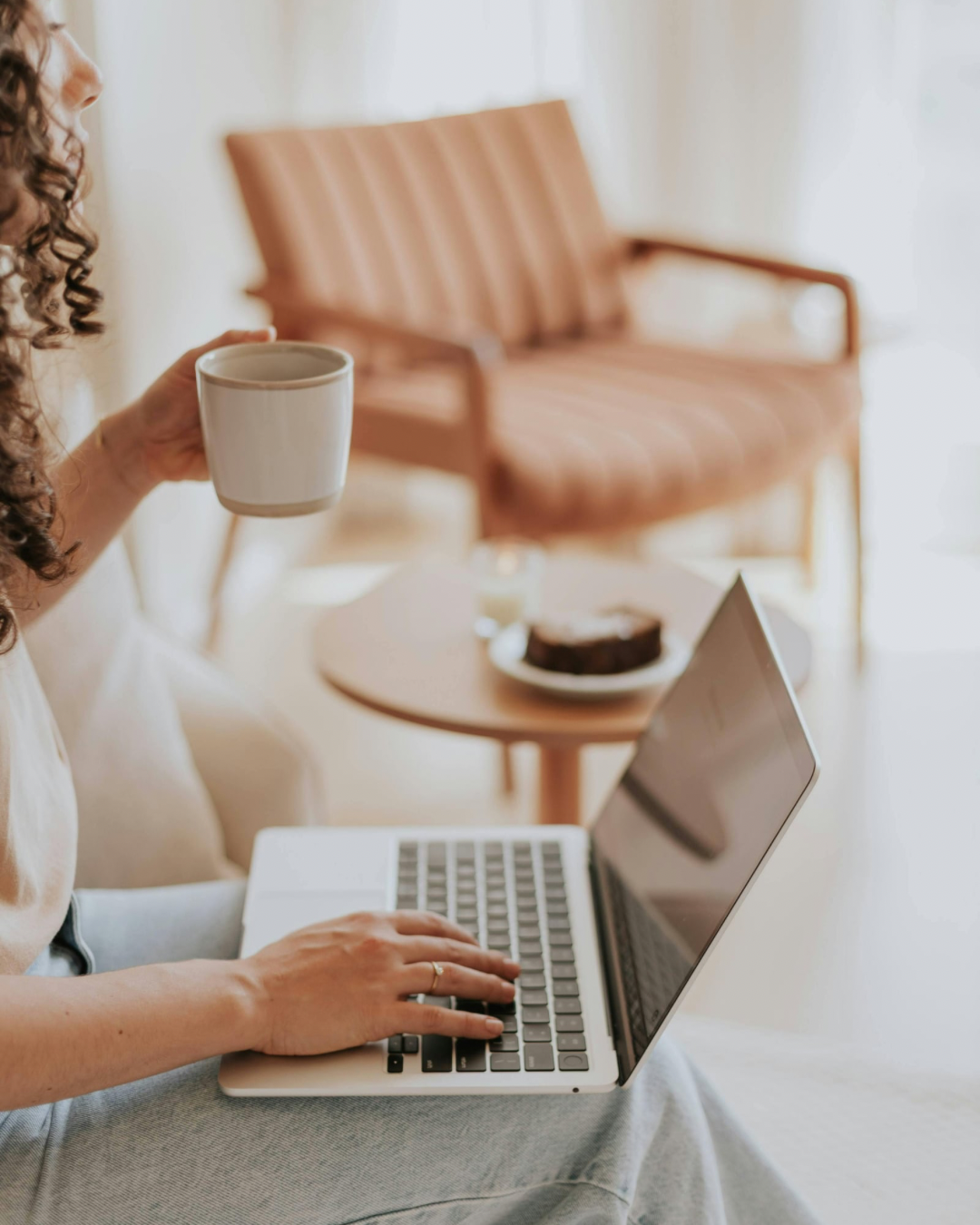 A woman with curly hair using a laptop, holding a coffee mug, in a cozy room with a pink sofa and a small round table with a dessert on it.