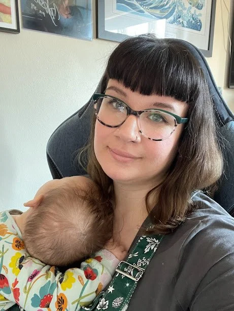 A woman with glasses and dark hair, smiling while holding a sleeping baby with a floral onesie, sitting in a chair in a room with framed artwork on the wall.