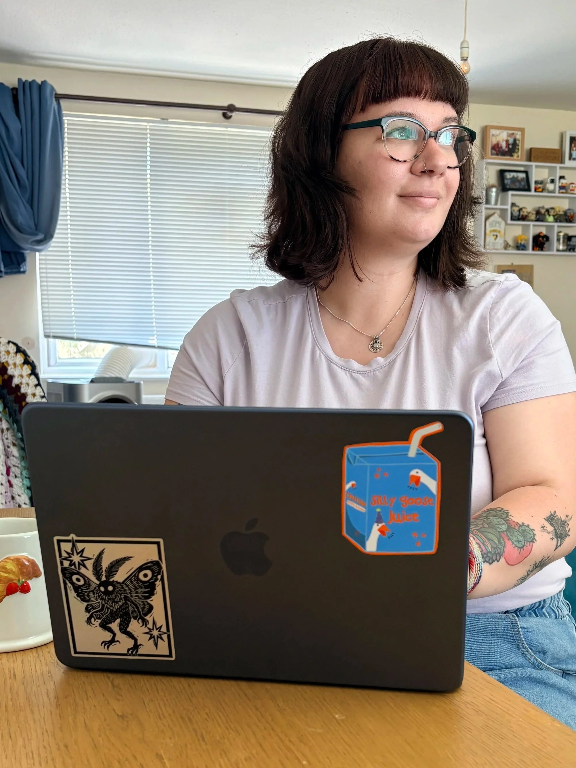 A woman with dark brown hair, glasses, and a nose ring, sitting at a table with a laptop that has stickers on it, including a cartoon owl and a box of salty goose juice juice. She has a colorful tattoo sleeve on her right arm. Behind her, there is a window with closed blinds, a bookshelf with various items, and a wall with framed photos.