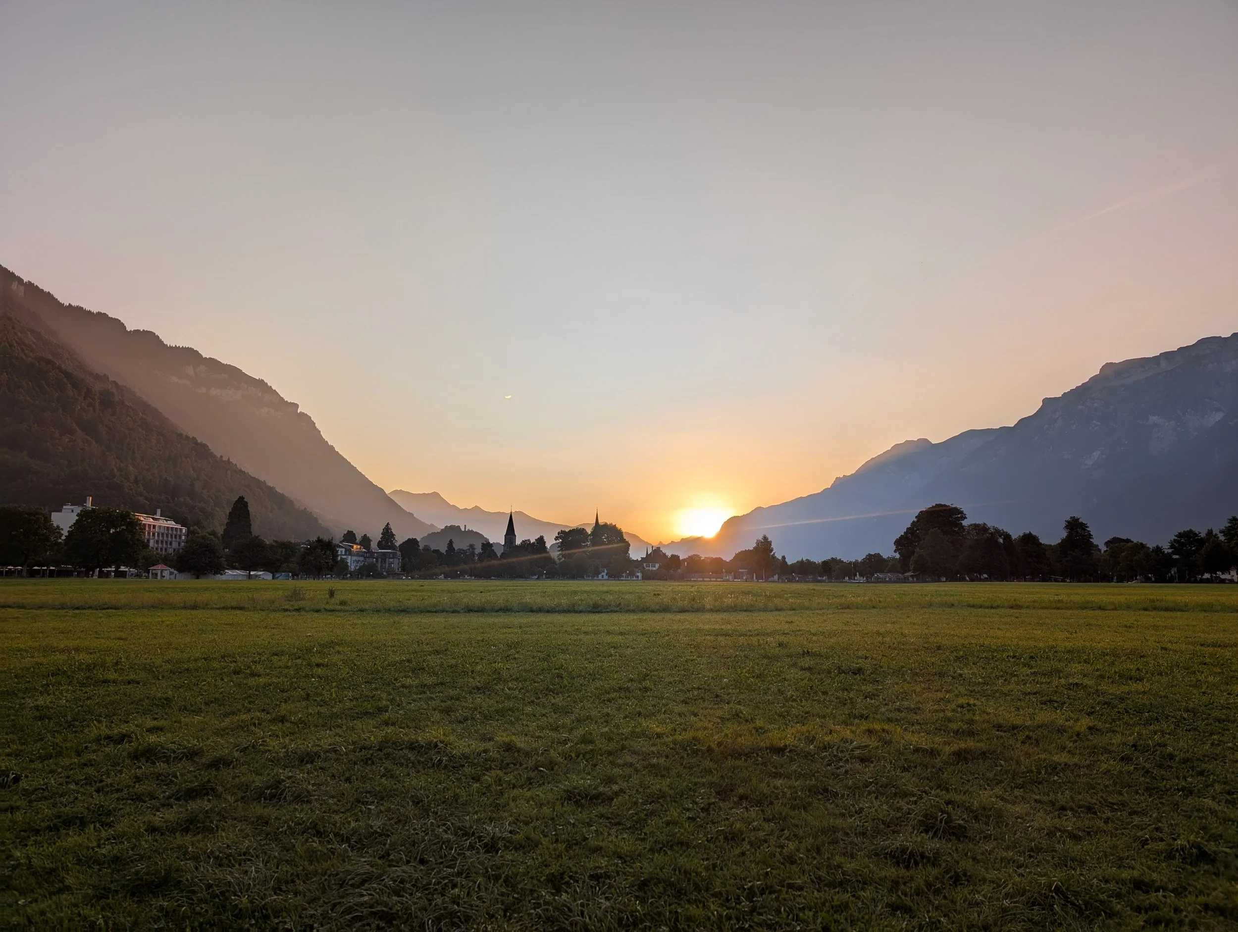 Sunset over mountains with a church steeple in the foreground and a grassy field.