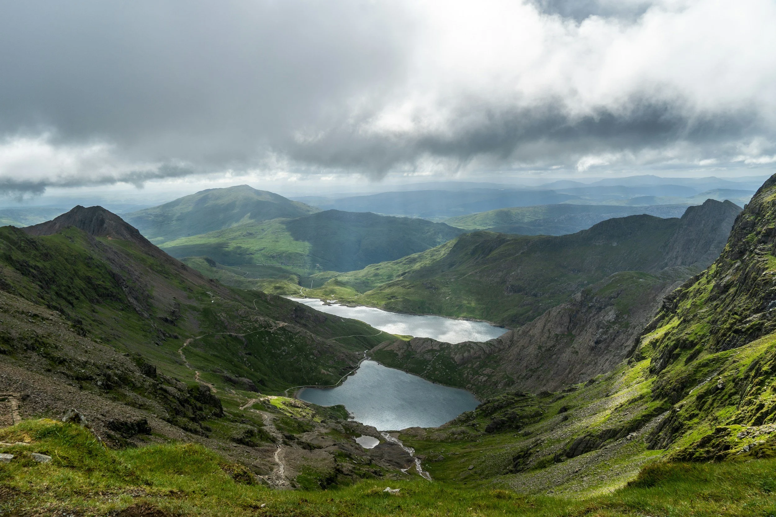 Mountain landscape with two lakes, green slopes, and cloudy sky.