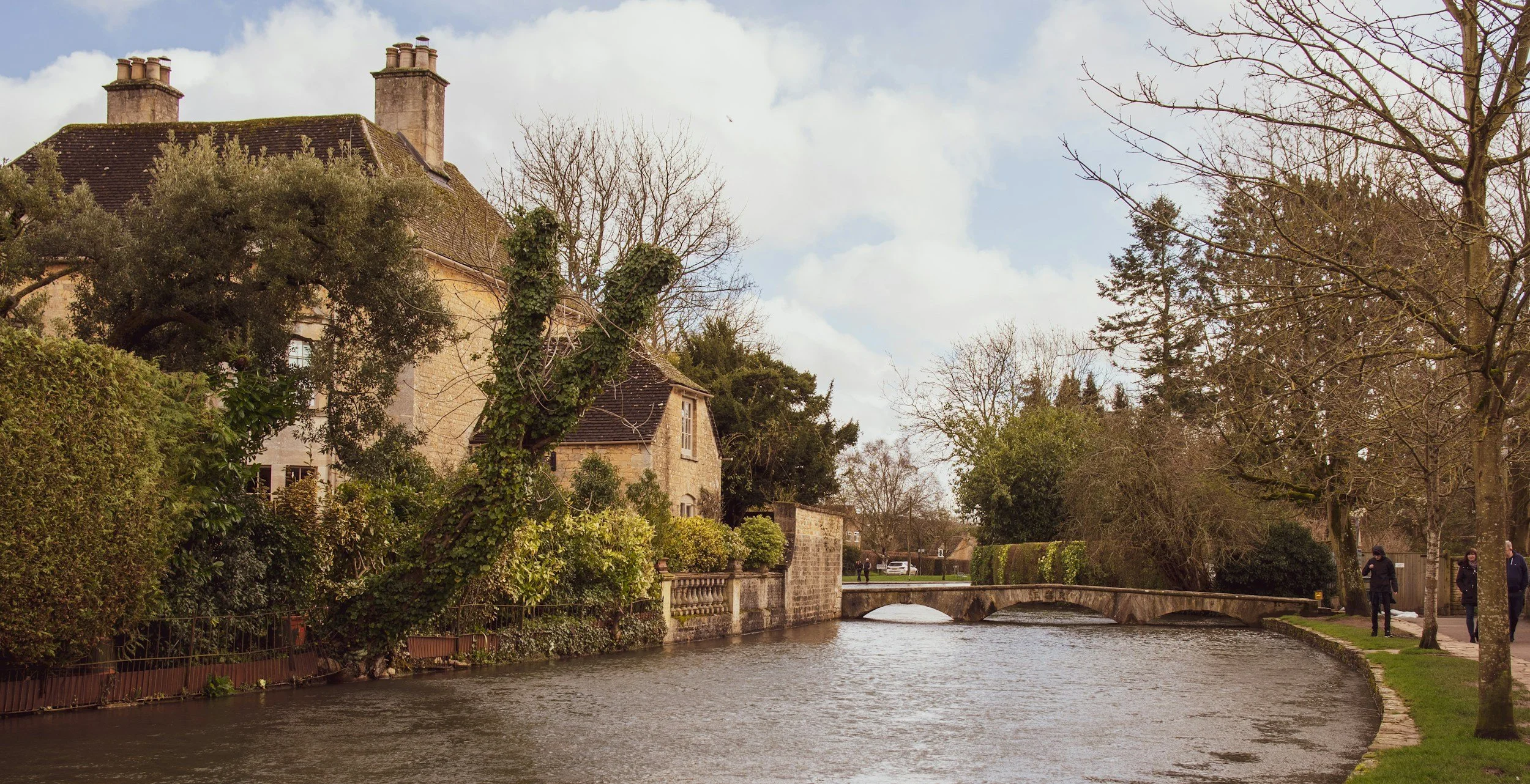 A peaceful riverside scene with a stone bridge, trees, a house, and pedestrians on the sidewalk.