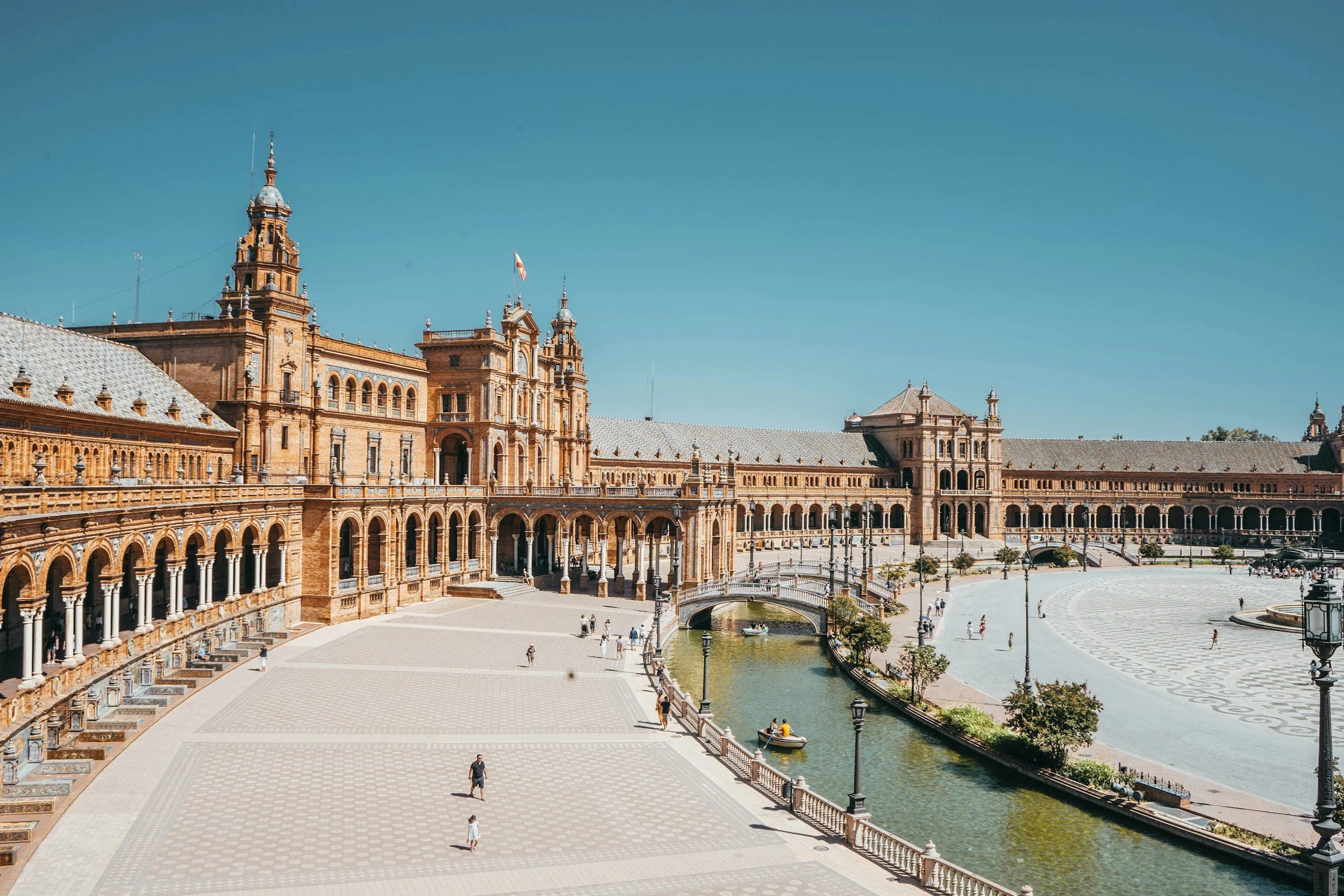 A large historic building with multiple towers and arches, a waterway running through a spacious square, and people walking and boating on a sunny day.