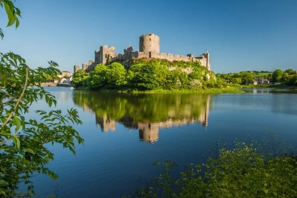 A castle on a small island surrounded by a river with green trees and clear blue sky reflected in the water.