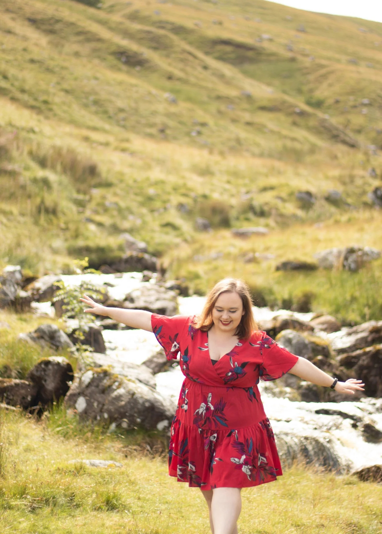 A woman in a red floral dress standing outdoors on a grassy area with arms outstretched and a joyful expression, with a mountain landscape in the background.