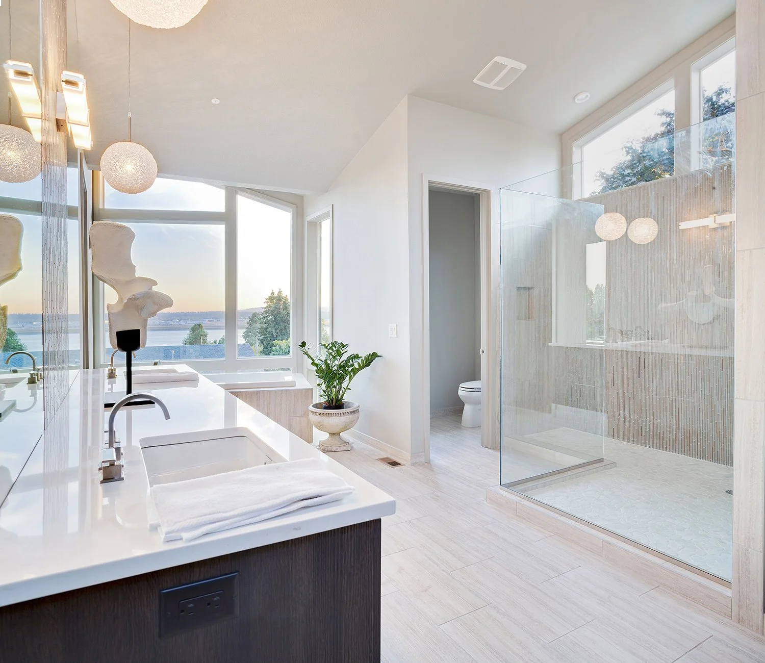 Modern bathroom featuring a double sink vanity, a potted plant, large windows with a view of trees and water, a toilet behind a door, and a glass-enclosed shower with beige tile walls.
