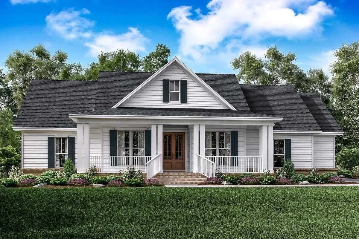 A white two-story house with a gabled roof, black shutters, front porch with white railings, and a wooden front door, surrounded by a landscaped yard with green grass and bushes.