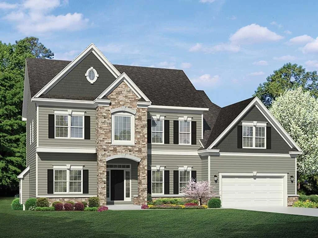 A large modern two-story house with gray and beige siding, black shutters, and a stone facade over the front entrance, surrounded by a well-maintained lawn and trees, under a blue sky with clouds.