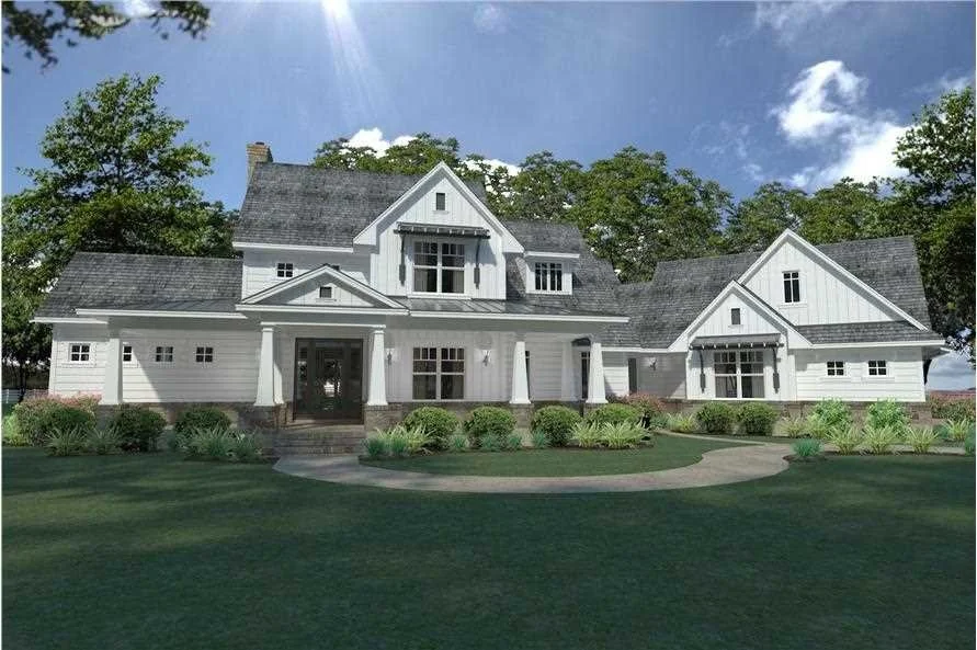 Large white two-story house with multiple gables, front porch, surrounded by green lawn and bushes, blue sky with clouds and trees in the background.