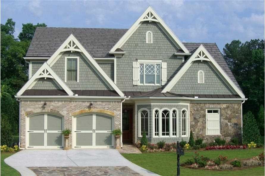 A large two-story house with a gray-green exterior, stone accents on the lower level, and a steeply pitched roof. It has a two-car garage, a front porch, and a prominent bay window. The front yard is landscaped with flowers and a trimmed lawn.