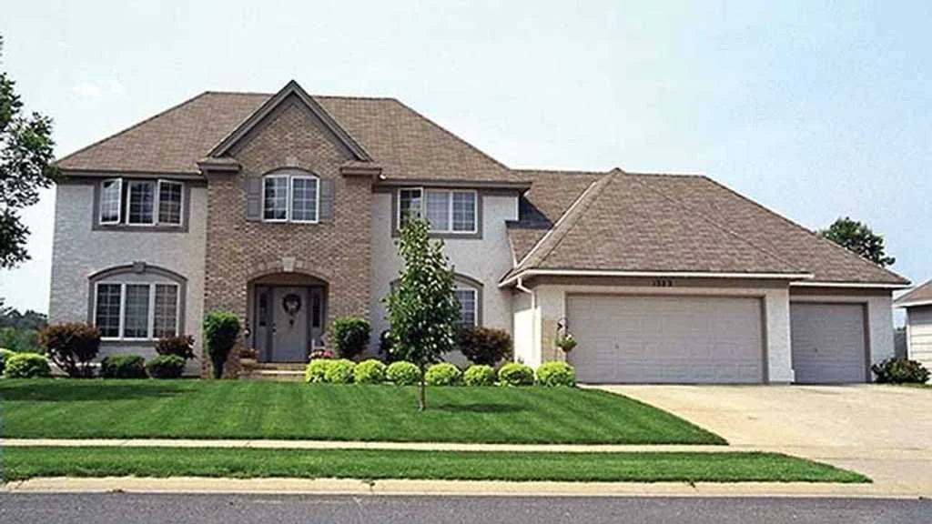 Two-story suburban house with brick and white siding, a three-car garage, and a manicured lawn with small bushes and a young tree.