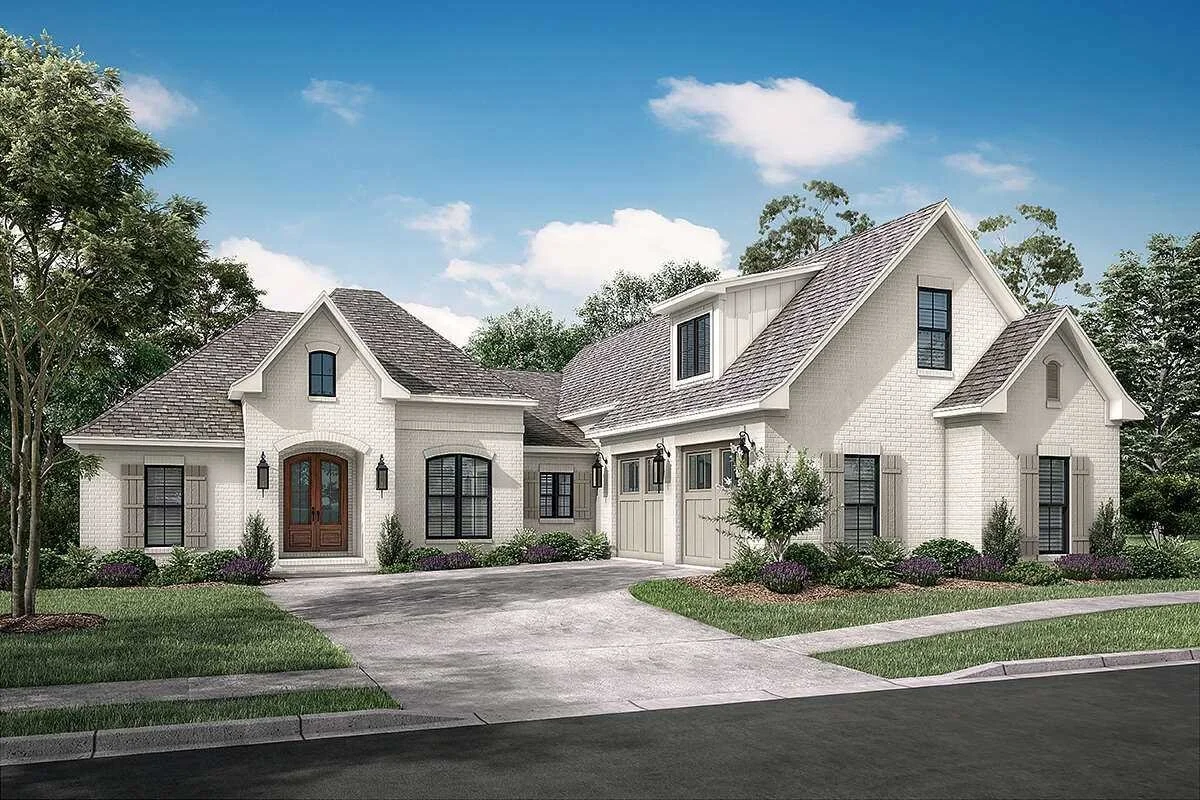 A large modern house with a white brick exterior, gabled roof, and two garage doors, surrounded by landscaped yard with shrubs and trees, under a blue sky with clouds.