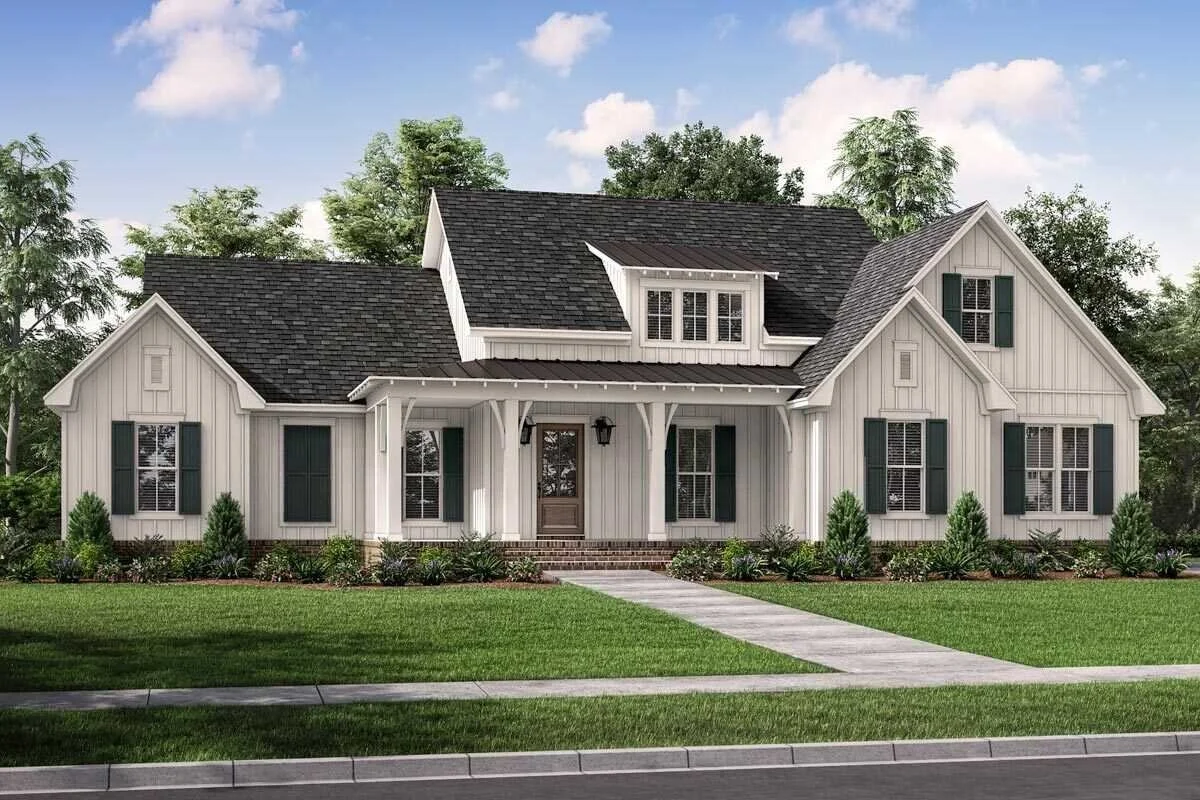 Front view of a large white house with a dark shingled roof, green shutters, a porch with white columns, and a walkway leading to the front door, surrounded by green grass and trees.