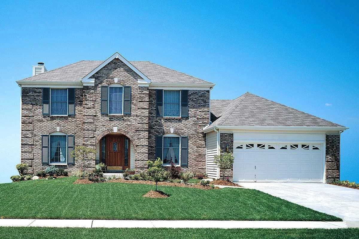 A two-story suburban house with brick exterior, gray shingle roof, and attached two-car garage, with a well-manicured lawn and small trees in front.