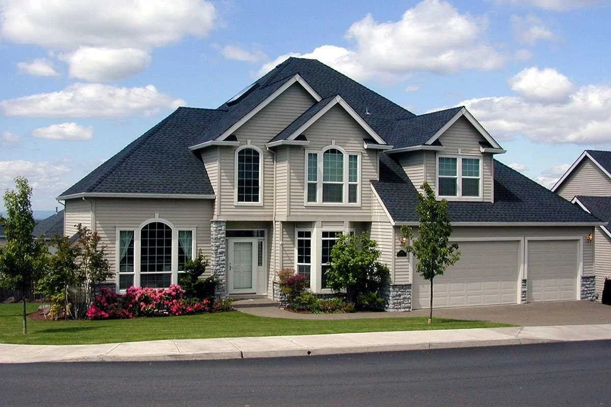 A two-story suburban house with beige siding, dark gray roof, multiple windows, and a two-car garage. There are trees and bushes in the front yard with a lawn and a sidewalk.