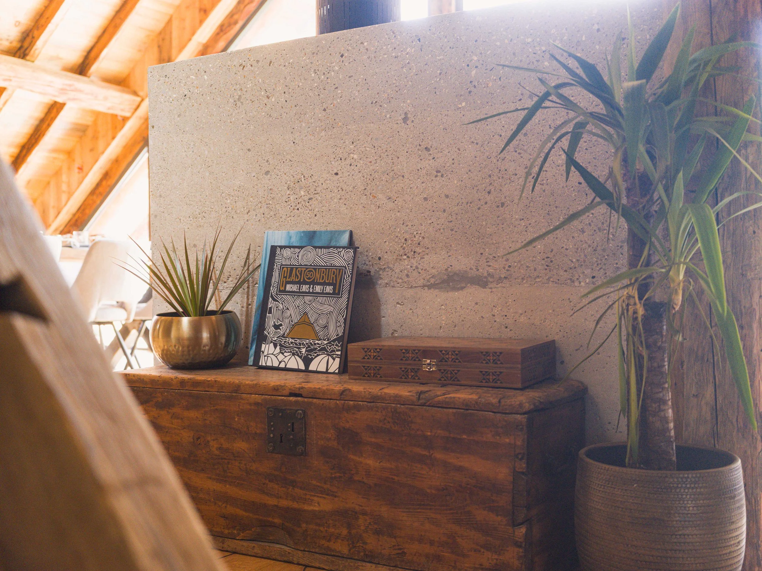 Vue intérieure d'un espace avec des poutres apparentes et un plafond vitré en pente. On y voit un grand mur en pierre beige mouchetée, une table en bois, une plante en pot et un plateau décoratif sur lequel repose un livre intitulé « Blastonbury ».