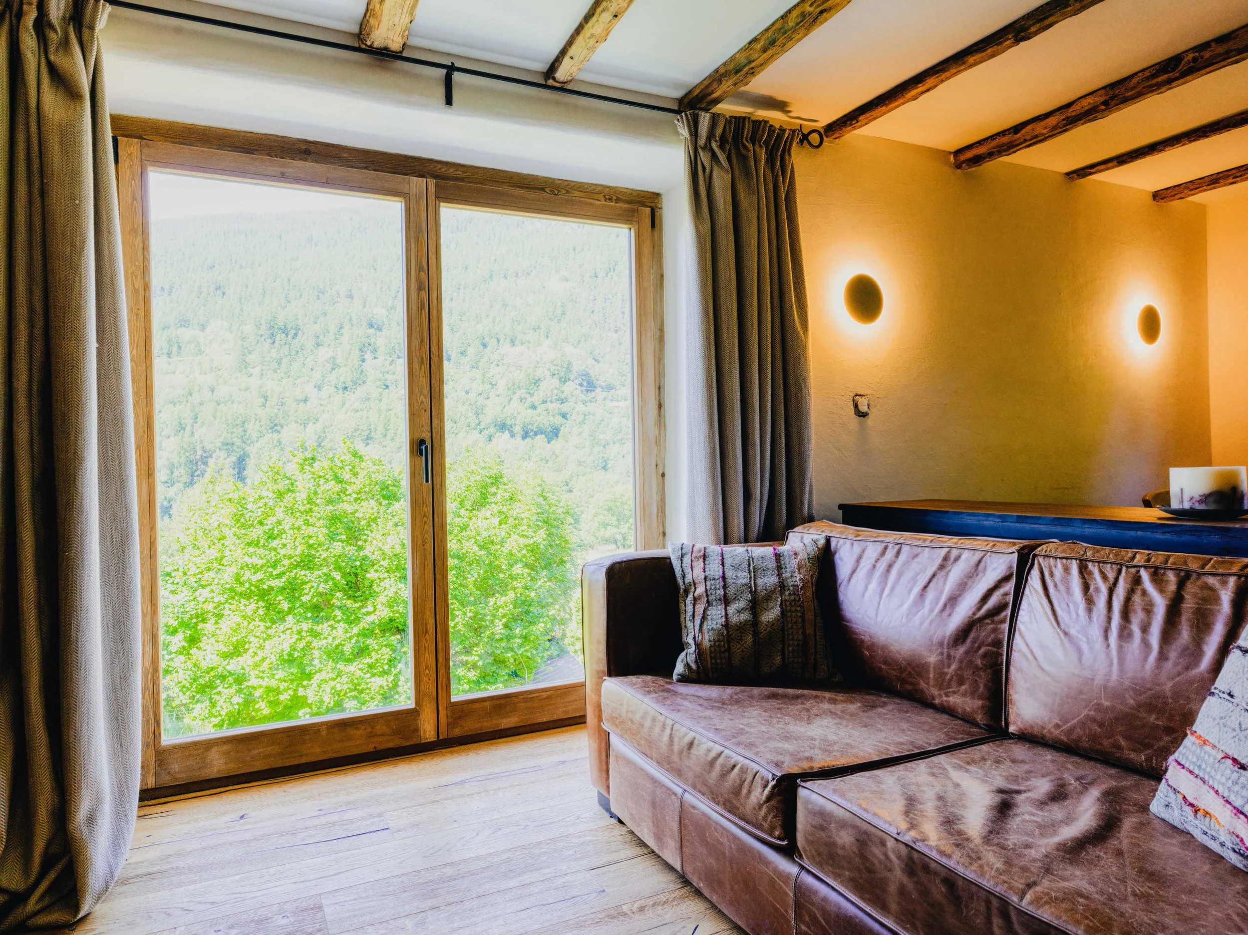 Living room with a large window showing a view of greenery and trees outside. The room has wooden flooring, beige curtains, a leather sofa, and modern wall-mounted round lights.