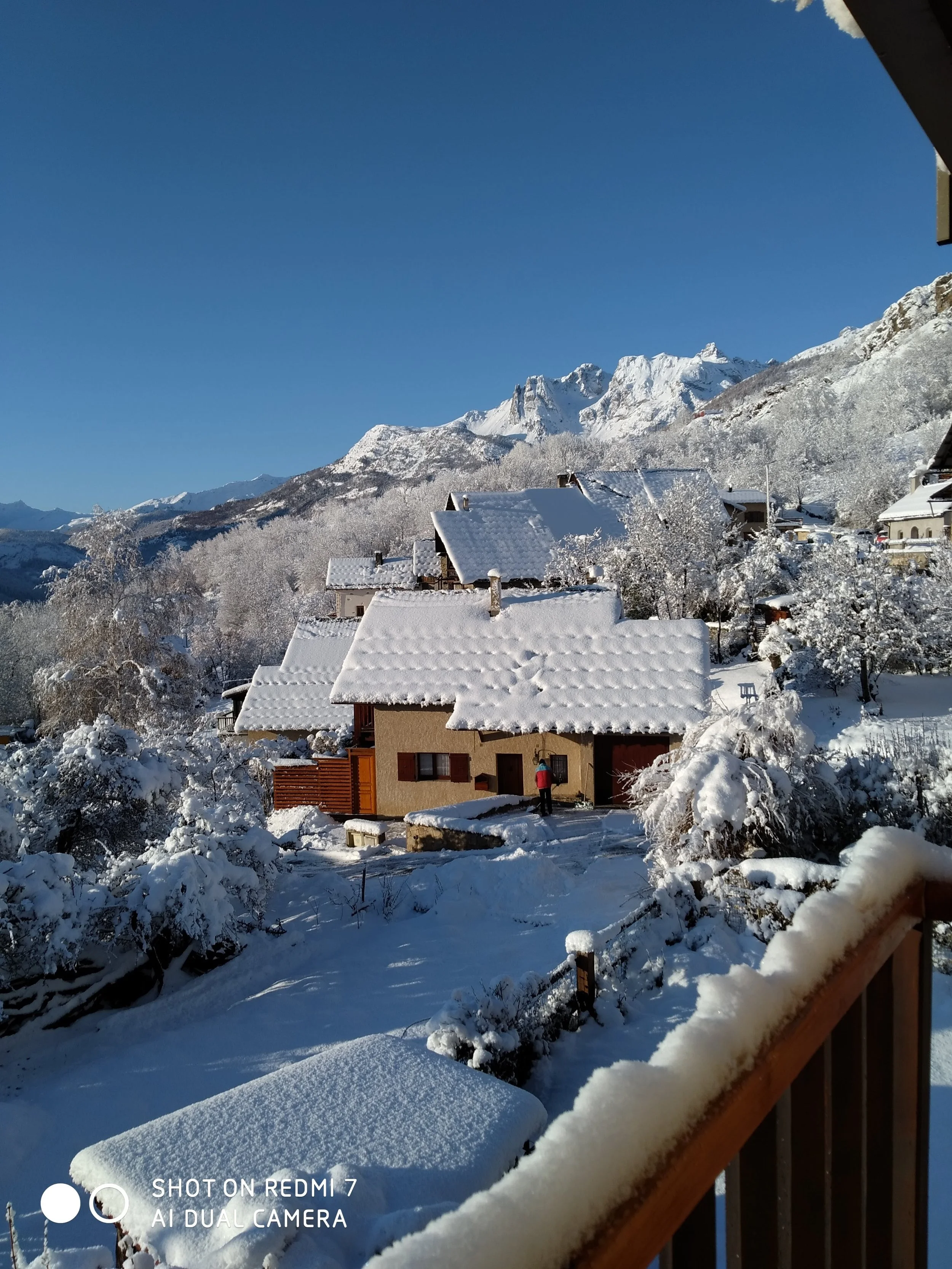 Snow-covered houses in a mountain village under a clear blue sky, with snow-laden trees and mountain peaks in the background.