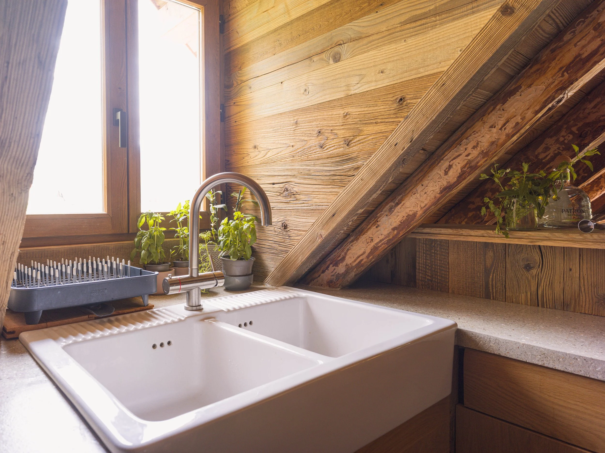 Une cuisine aux murs en bois, avec une fenêtre laissant entrer la lumière naturelle, un évier blanc, un robinet et des plantes en pot sur le rebord de la fenêtre.
