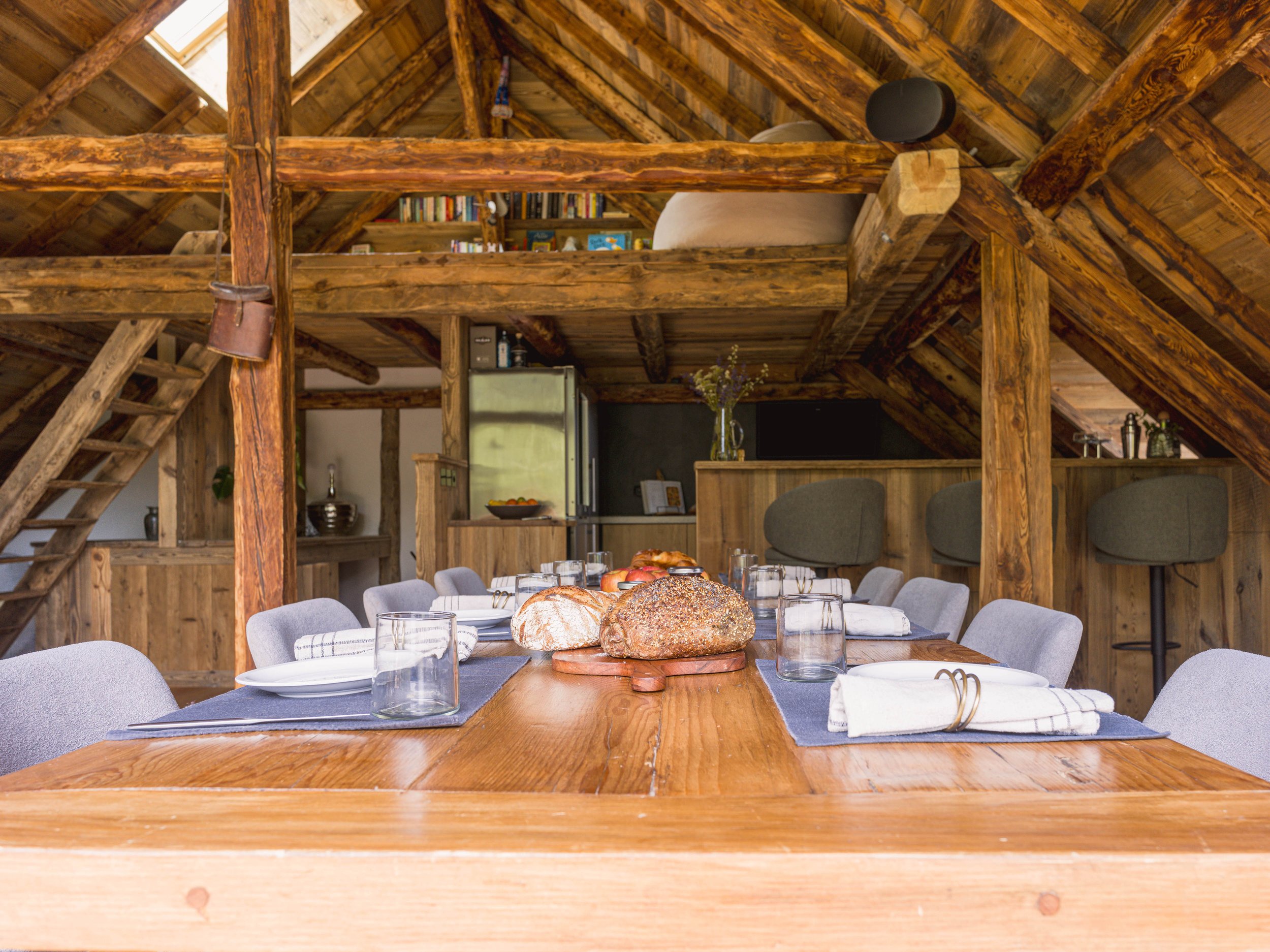 Une salle à manger dans un chalet rustique, avec une longue table en bois dressée avec du pain, des verres et des serviettes, entourée de chaises rembourrées bleues. À l'arrière-plan, on aperçoit une mezzanine avec des étagères, un escalier et un coin cuisine équipé d'un réfrigérateur et de divers