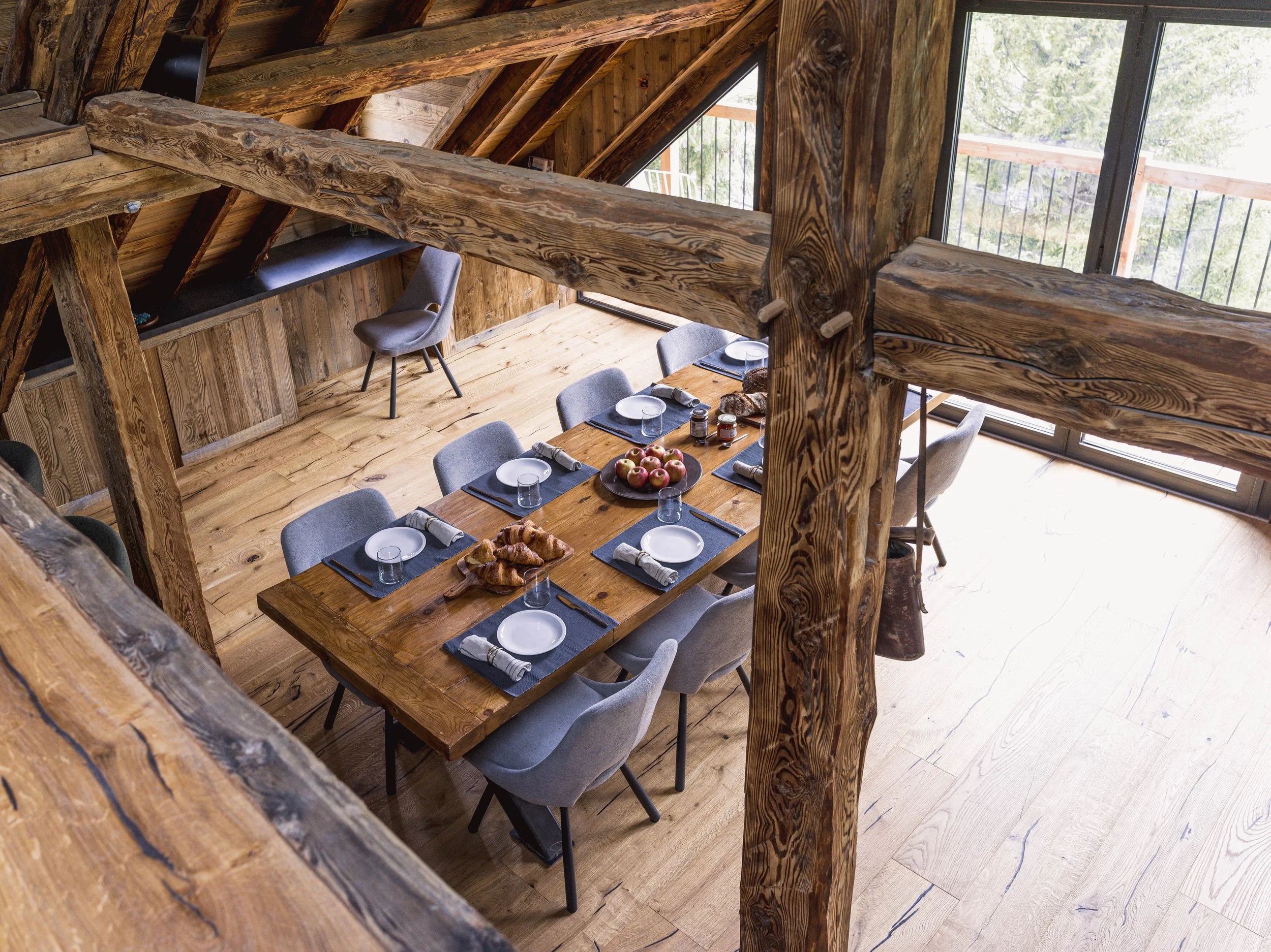 Une salle à manger rustique avec une longue table en bois dressée pour le repas, avec des assiettes, des verres et un bol de pommes, entourée de chaises rembourrées grises. La pièce est dotée de poutres apparentes et de grandes fenêtres donnant sur les arbres à l'extérieur, baignées de lumière naturelle.