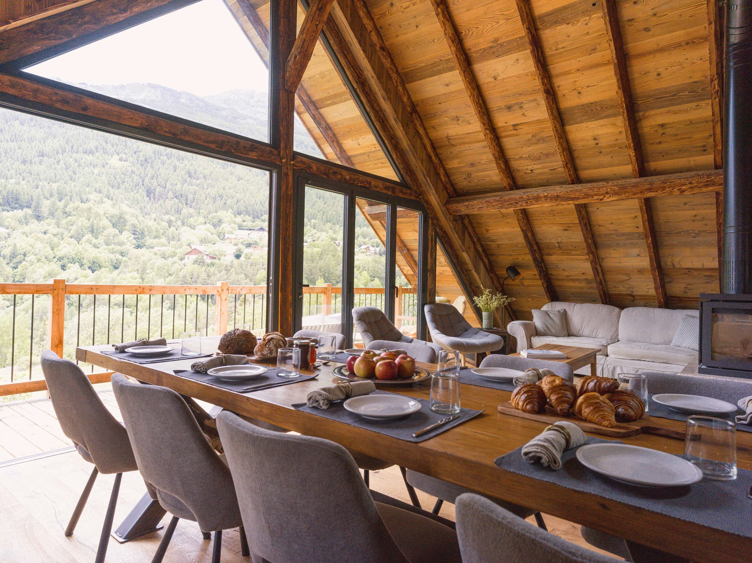 Vue intérieure d'une cabane en bois avec un haut plafond en pente et de grandes fenêtres donnant sur un paysage montagneux. Une table à manger dressée pour le petit-déjeuner, avec des assiettes, des verres et du pain, entourée de chaises rembourrées grises. À l'arrière-plan, un coin salon confortable