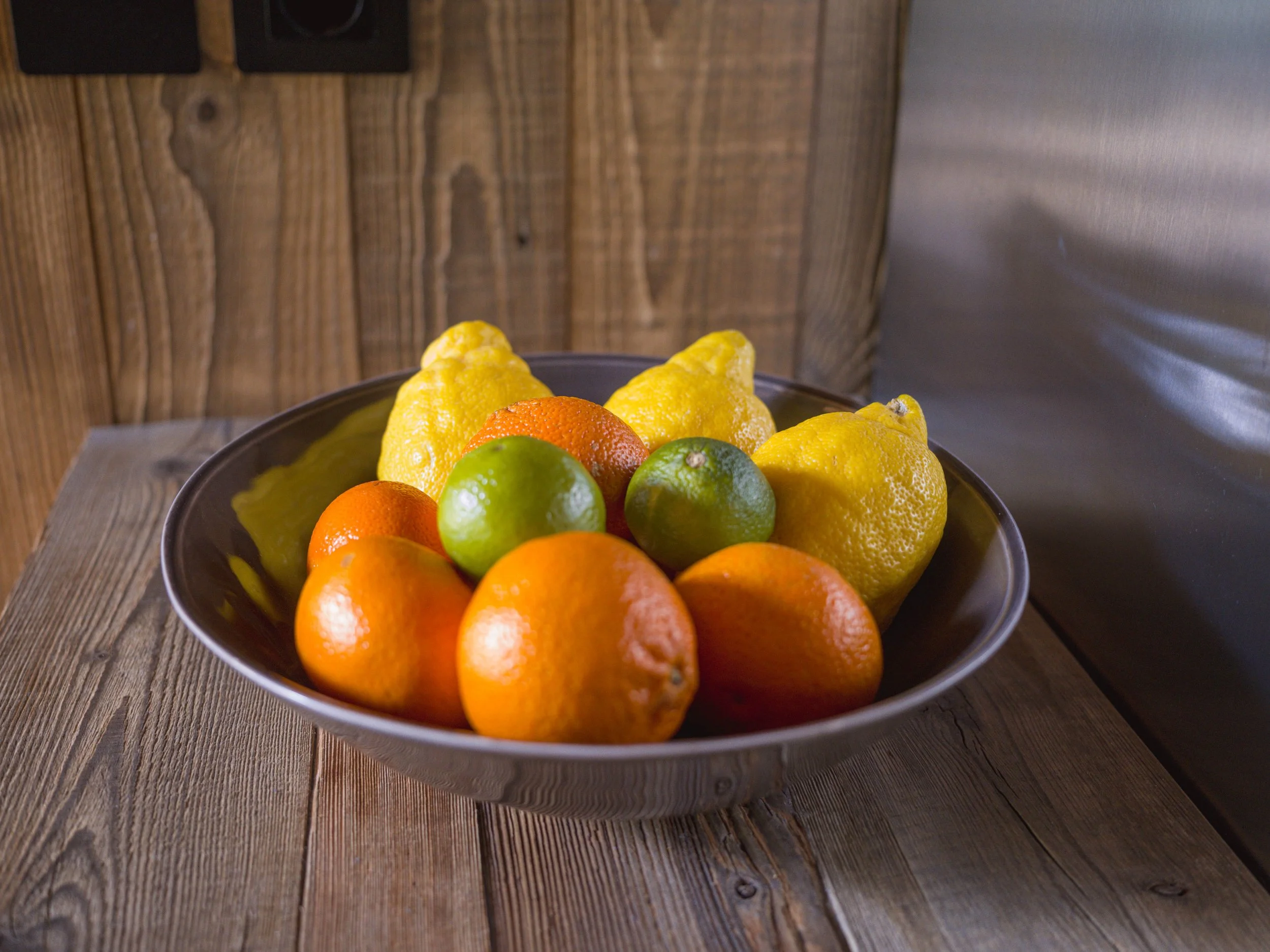Un bol noir rempli d'un assortiment d'agrumes, notamment des oranges, des citrons, des citrons verts et des mandarines, posé sur une table en bois avec, en arrière-plan, un mur en bois rustique.