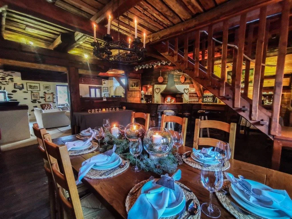 A rustic dining room set for a festive holiday meal with a decorated table, candles, and wooden chairs, inside a cozy cabin with wood-paneled walls and a staircase leading upstairs.
