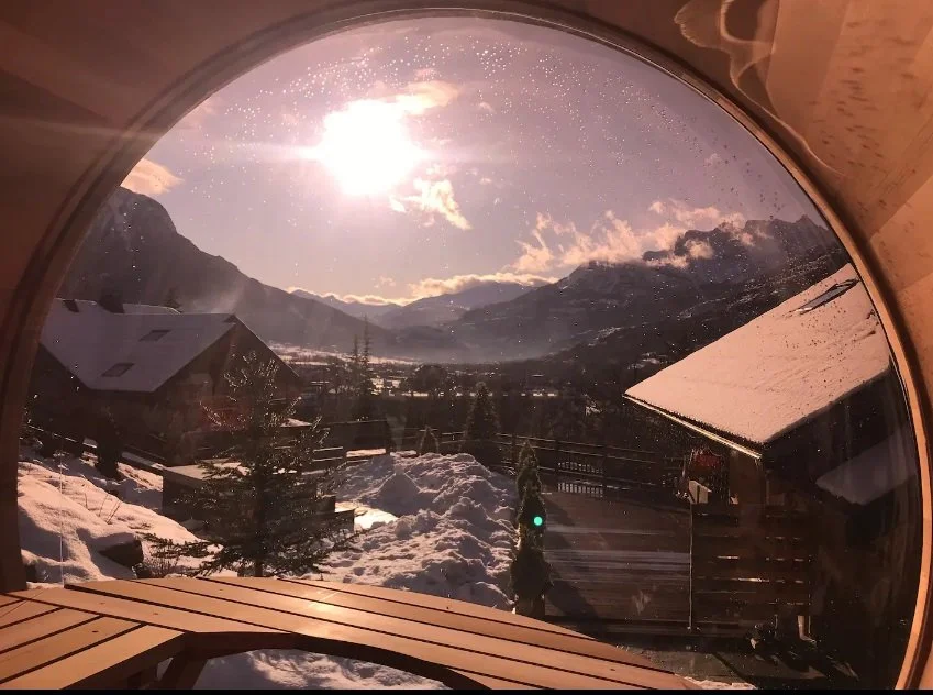 View of snowy mountains and houses through a circular window with sunlight and clouds.