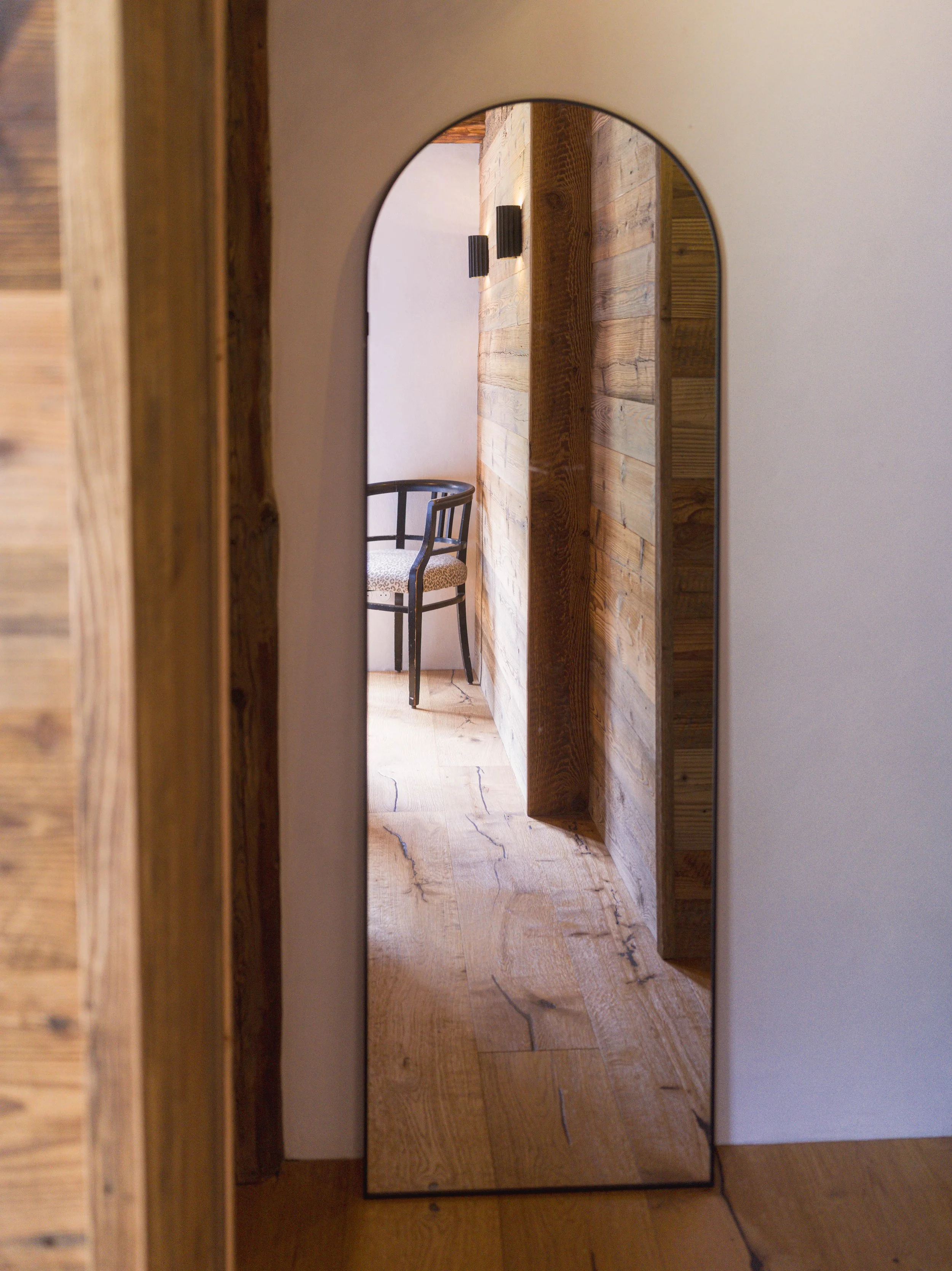 Un miroir en pied appuyé contre un mur en bois, avec une chaise visible à proximité à travers l'embrasure d'une porte.