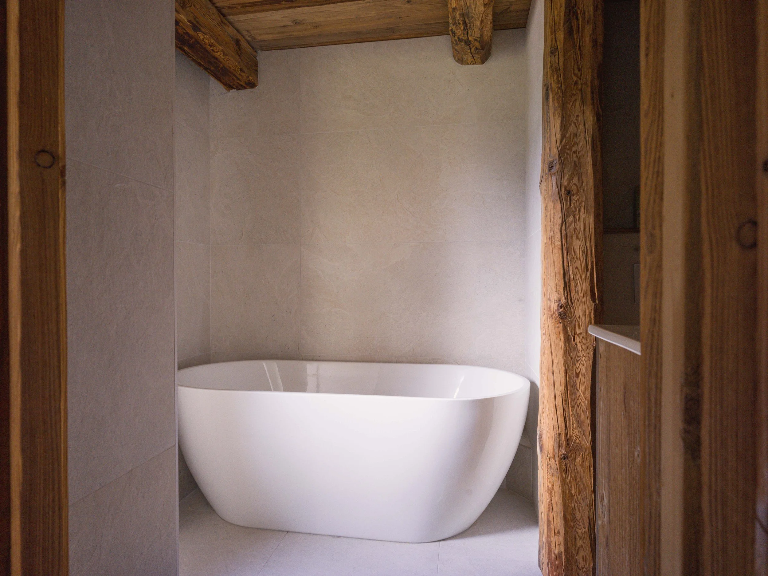 Une baignoire blanche dans une petite salle de bains aux murs carrelés de beige et au plafond en bois. La photo a été prise depuis l'embrasure de la porte, en regardant vers l'intérieur de la pièce.