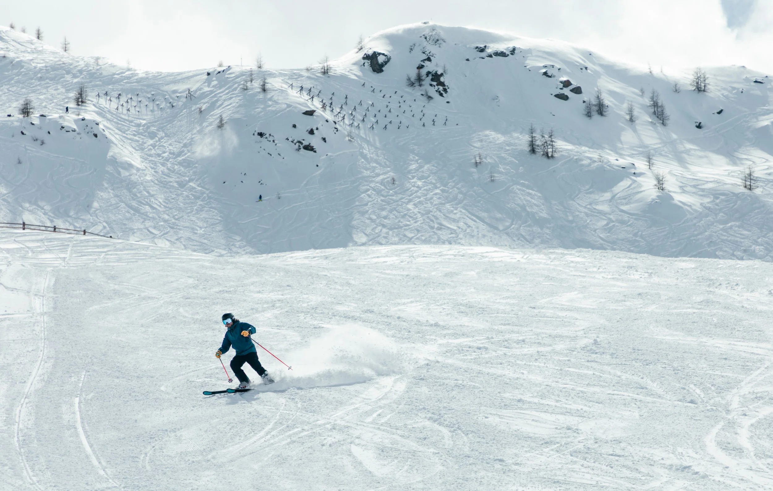 A skier in blue gear glides down a snow-covered slope with a mountain and sparse trees in the background.