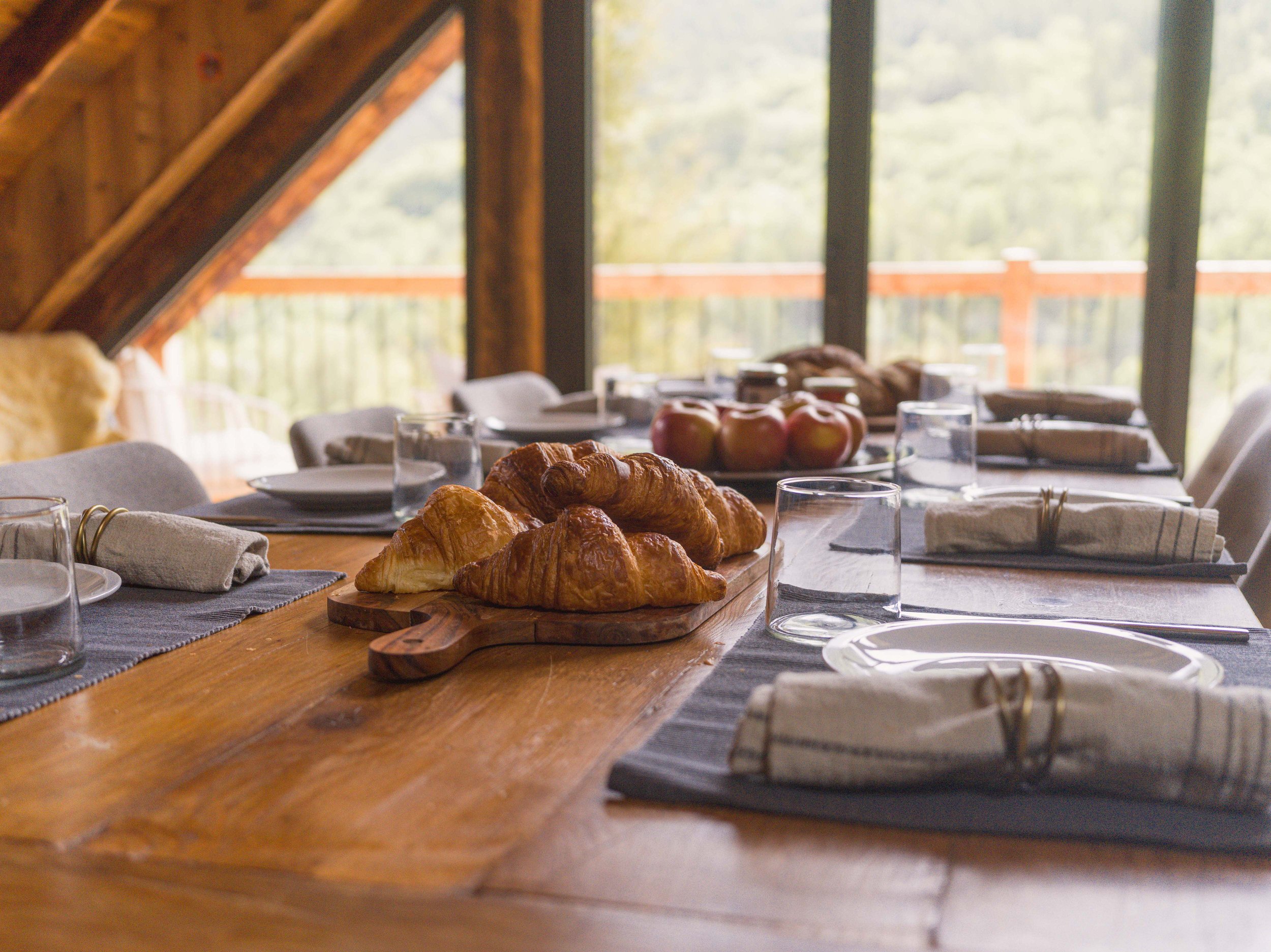 Une table à manger dressée avec des croissants, des pommes, des verres et des serviettes, devant une grande fenêtre donnant sur des arbres.