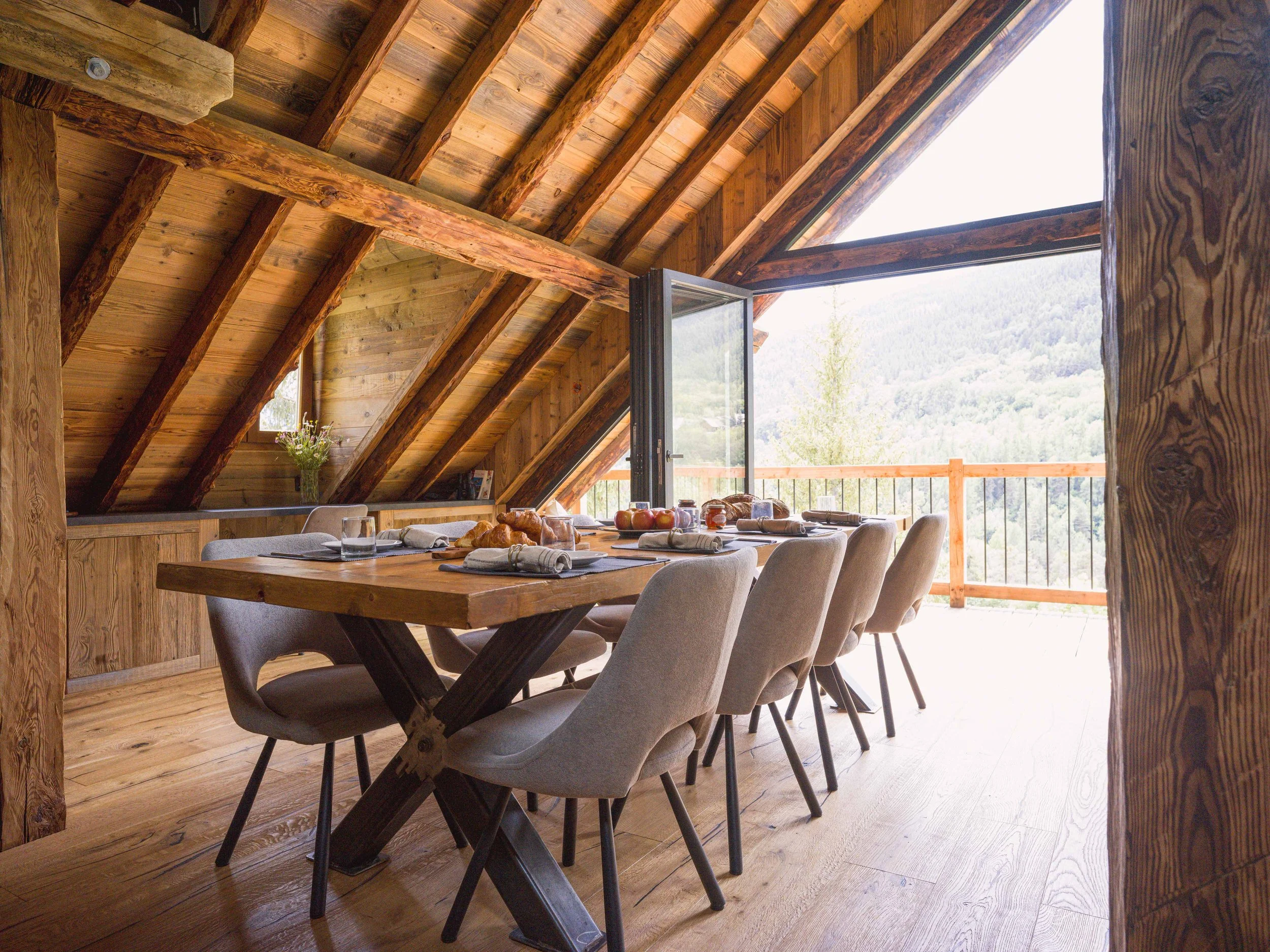 Une salle à manger avec une grande table en bois dressée de sets de table, de couverts et de pain, entourée de chaises rembourrées, le tout dans un intérieur rustique en bois doté de grandes fenêtres offrant une vue imprenable sur les montagnes.