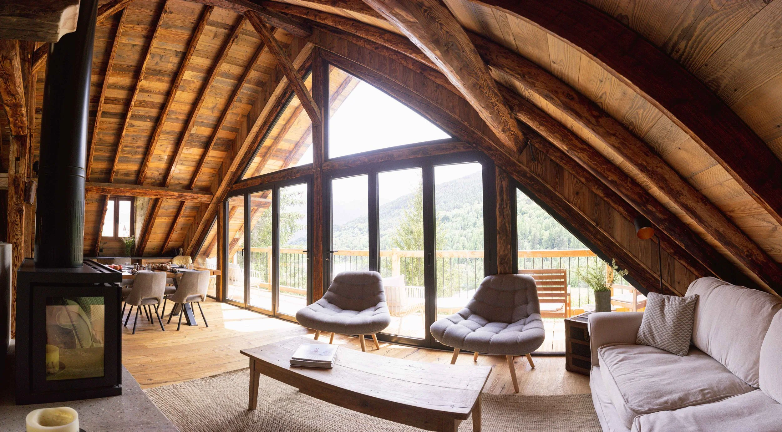 Salon avec plafond voûté en bois, grandes baies vitrées donnant sur un balcon extérieur, deux fauteuils gris, un canapé blanc et une vue sur les montagnes.