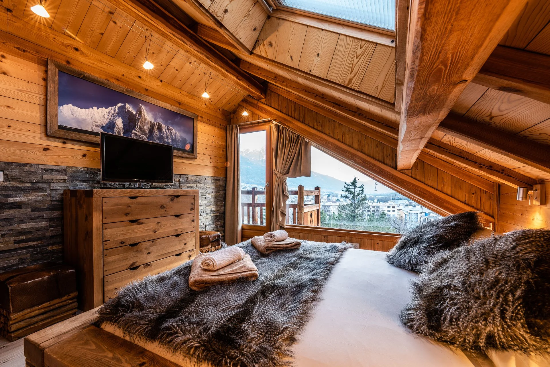 Cozy mountain cabin bedroom with wooden walls and sloped ceiling, large window showing mountain view, bed with fur throw and pillows, wall-mounted TV, framed mountain photo, skylight, and candles.