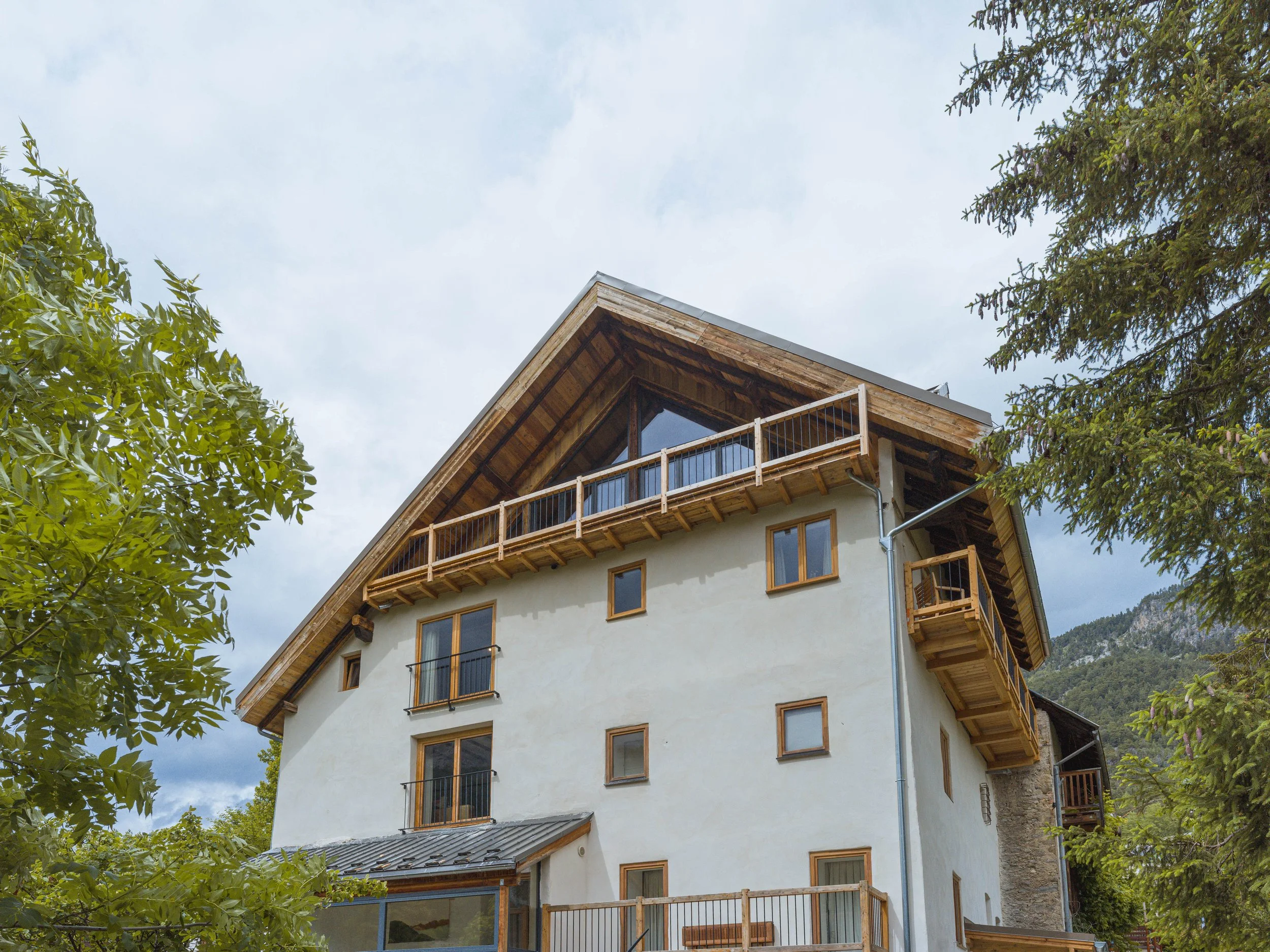 A modern multi-story house with white exterior walls, wooden window frames, and a balcony with a glass railing, surrounded by green trees, set against a partly cloudy sky.