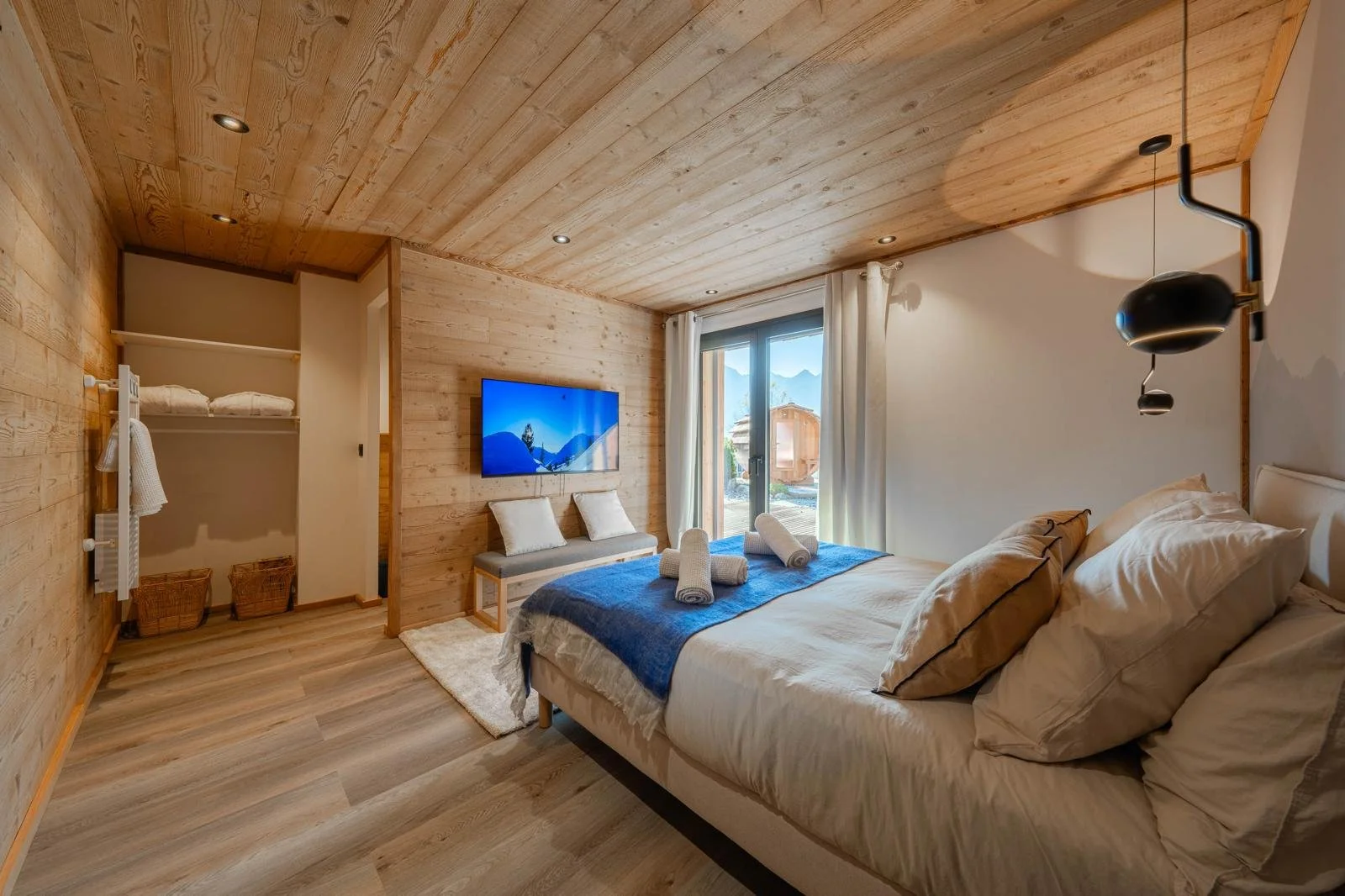 A cozy bedroom with wooden walls and ceiling, a large bed with beige bedding and pillows, a TV on the wall, a sliding glass door with white curtains leading outside, and a bench underneath the TV with cushions and towels, featuring a small closet are