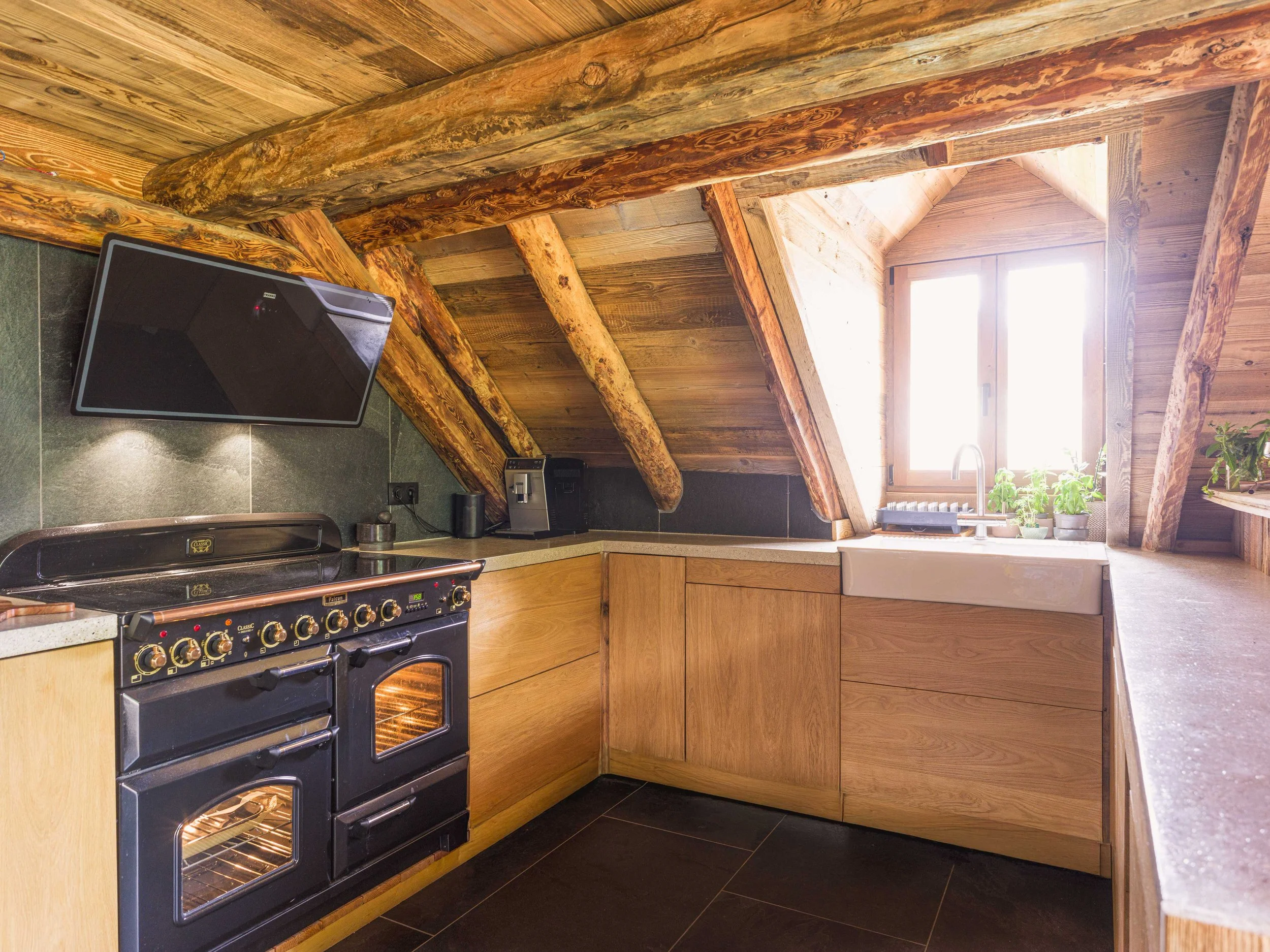 Une cuisine avec des rondins de bois et des lambris aux murs et au plafond, une cuisinière noire aux boutons dorés, une fenêtre baignée de soleil, un évier avec des plantes vertes à proximité et un sol carrelé noir.