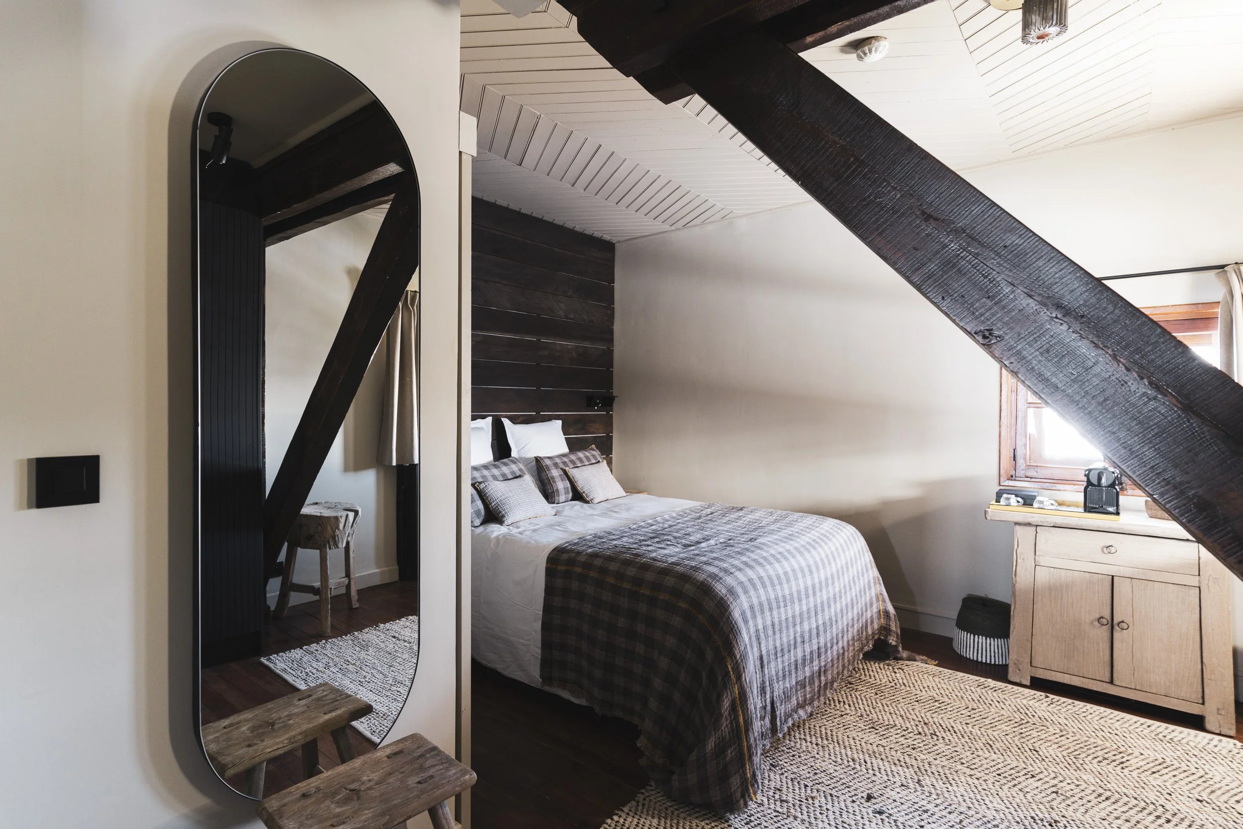 A cozy bedroom with a sloped wooden ceiling, a bed with plaid blankets, an arched mirror, a wooden dresser, and a window with natural light.