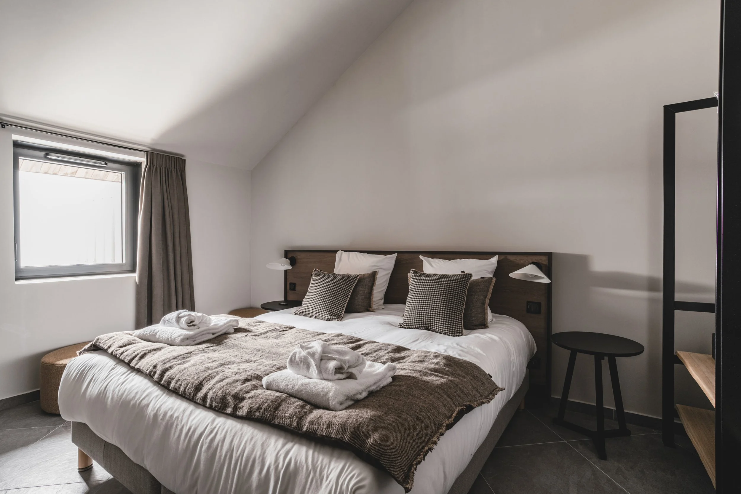 Modern bedroom with a large bed featuring white sheets, multiple pillows, and a brown blanket. Wooden headboard with attached reading lamps on each side. Towels folded into swan shapes on the bed. A window with gray curtains, black side tables, and m