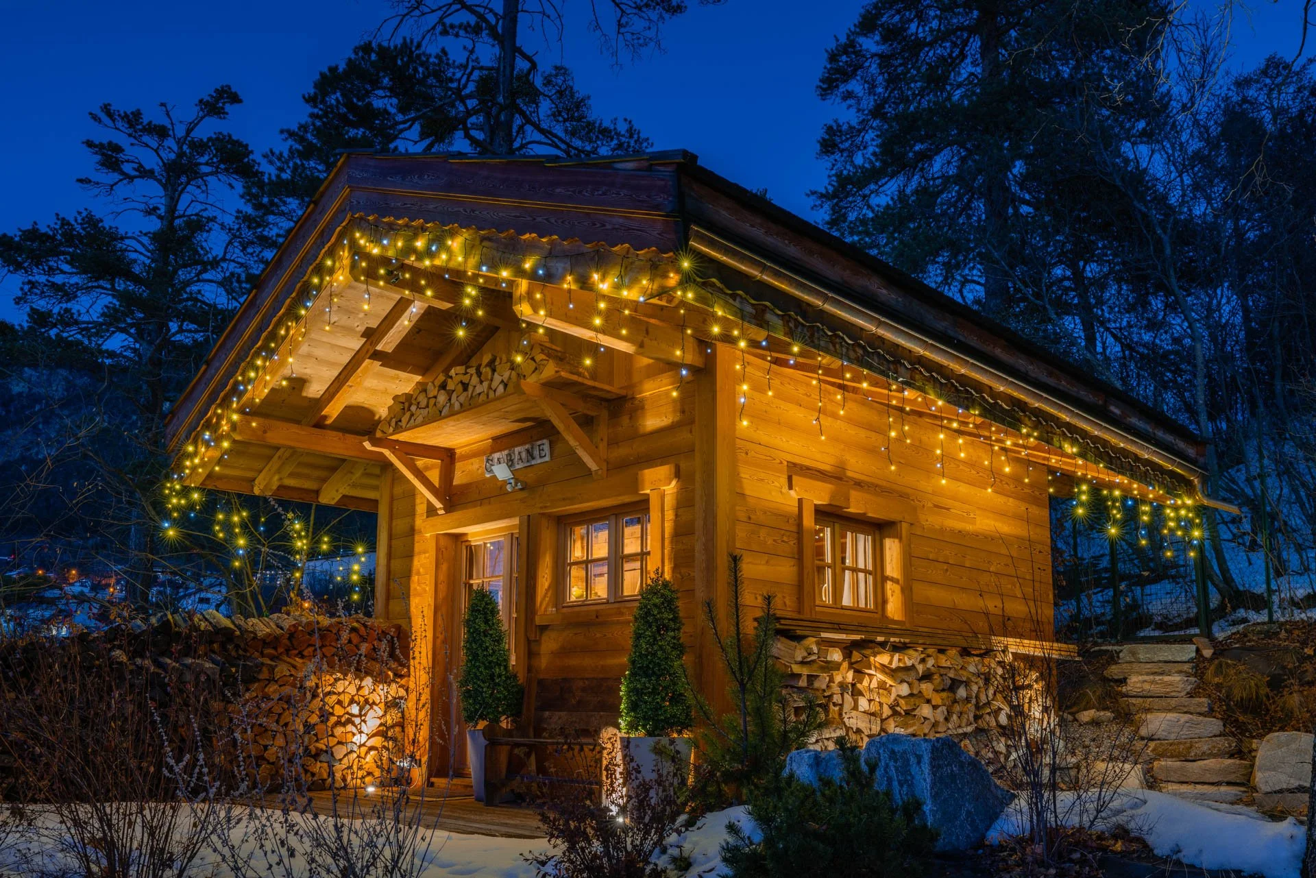 A cozy wooden cabin decorated with string lights at night, with forest trees in the background and snow on the ground.