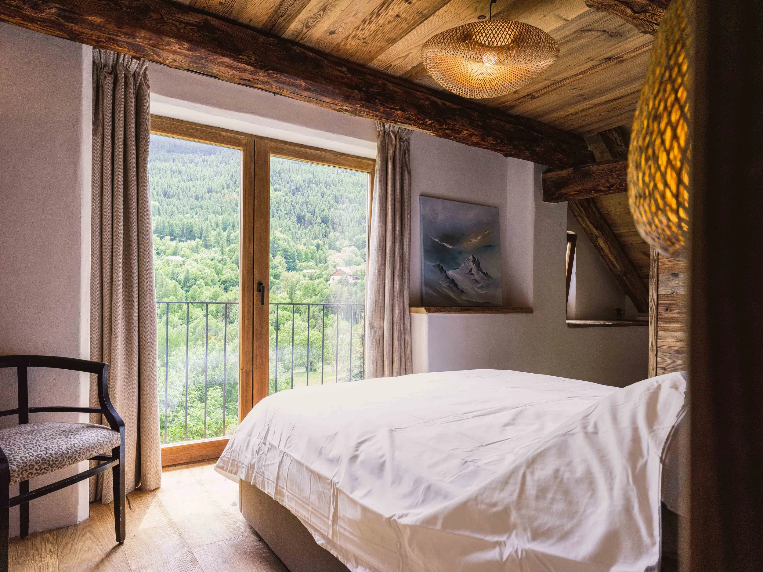 Une chambre chaleureuse dotée d'une grande fenêtre donnant sur un paysage verdoyant et montagneux. La pièce présente des poutres apparentes, des rideaux aux tons neutres, un lit blanc recouvert d'un couvre-lit sobre et un tableau représentant des montagnes accroché au mur. Une chaise avec un coussin à motifs