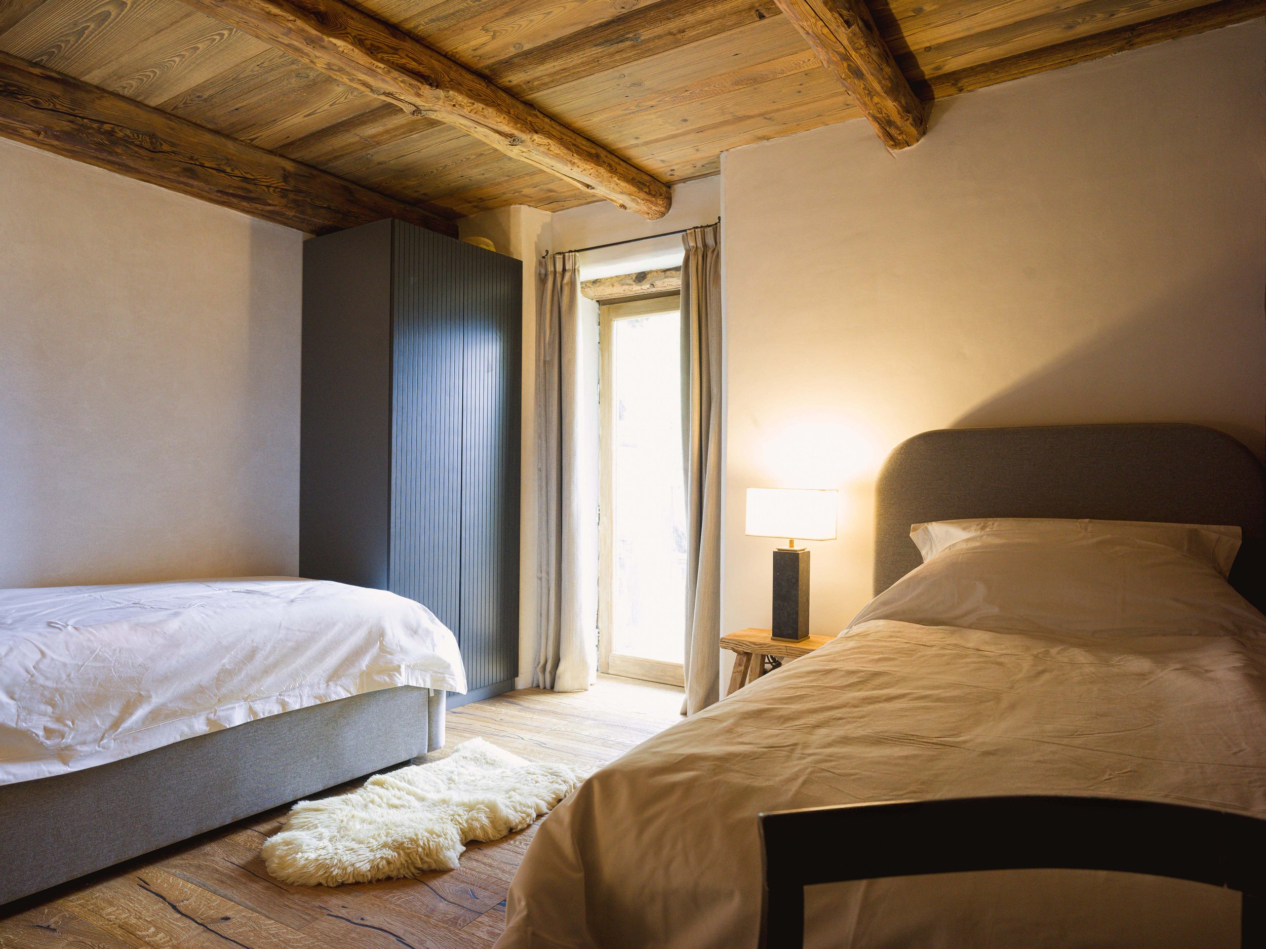 Deux lits recouverts de draps beiges et blancs dans une chambre rustique dotée de poutres apparentes, d'une petite table de chevet avec une lampe, d'une armoire noire, d'une fenêtre avec des rideaux et d'un tapis blanc moelleux.