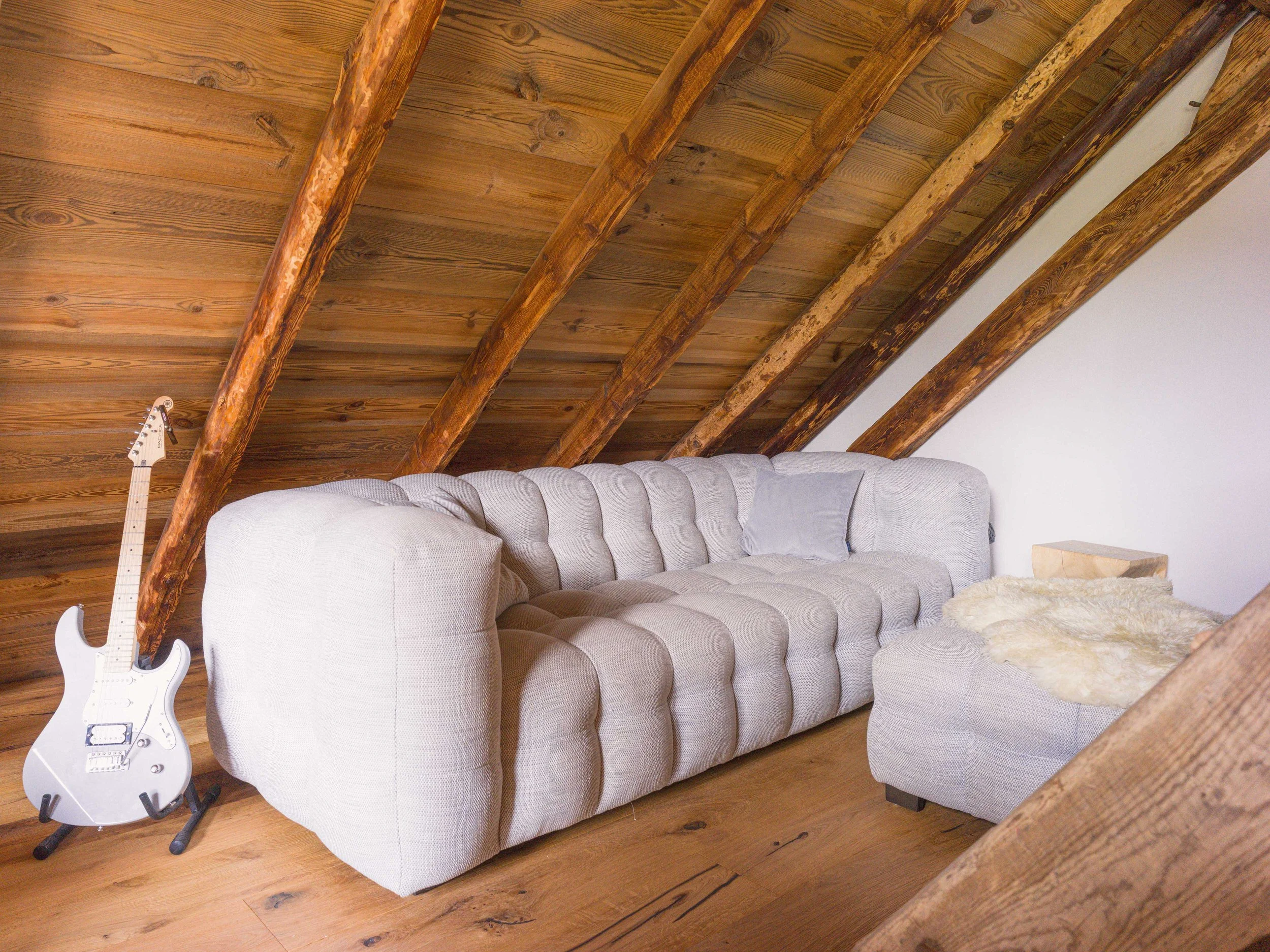 Un espace de vie chaleureux aménagé dans les combles, avec un plafond en pente en bois soutenu par des poutres rustiques. Une guitare électrique blanche posée sur un support se trouve à côté d'un canapé rembourré de couleur claire, et un tapis en peau de mouton recouvre le parquet.