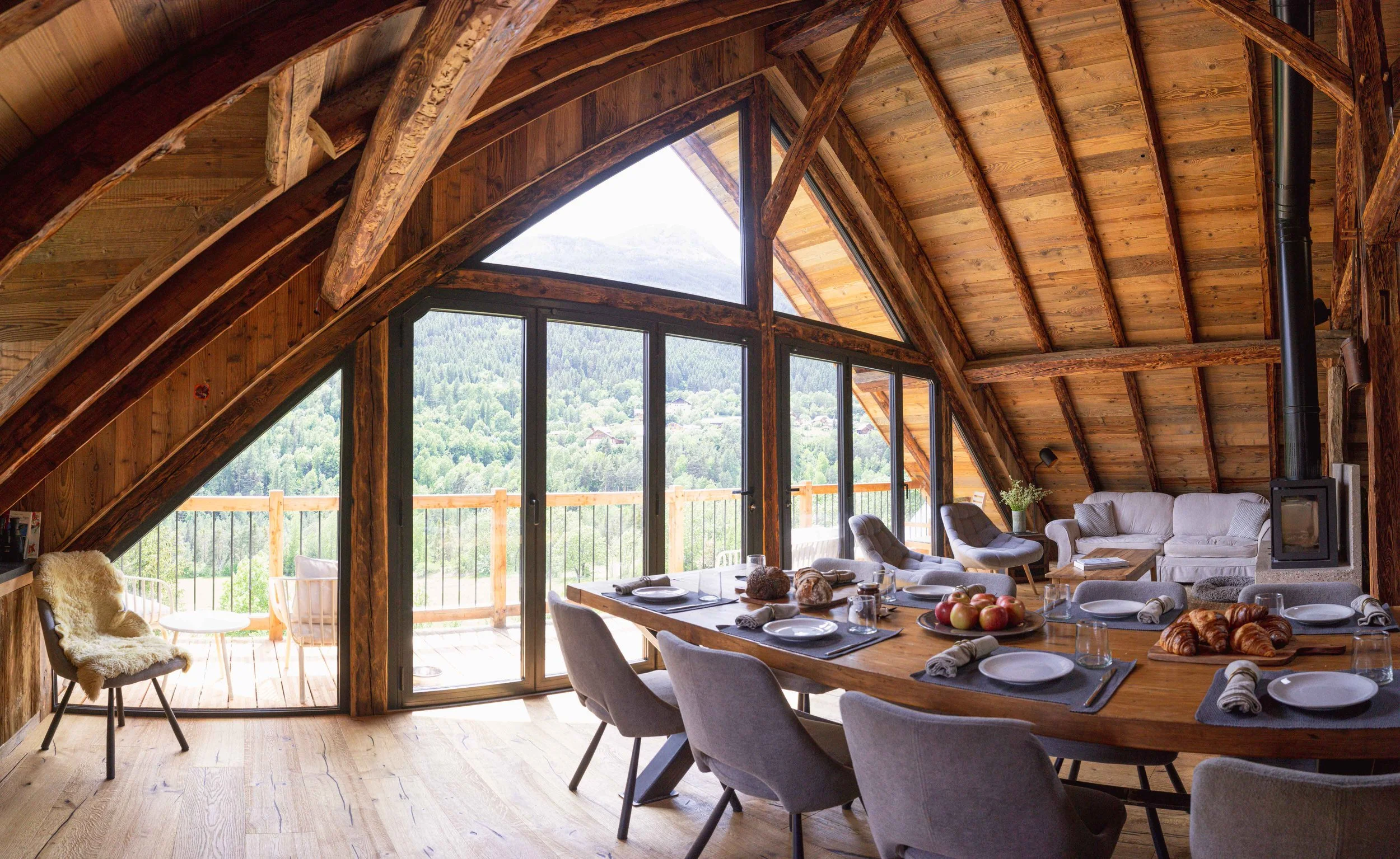 Intérieur d'un chalet de montagne en bois avec un haut plafond voûté, de grandes fenêtres offrant une vue panoramique sur les montagnes, une table à manger dressée pour le repas et un coin salon confortable avec des chaises et un canapé.