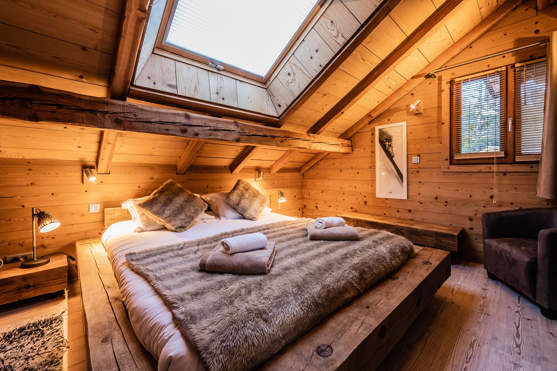 Cozy bedroom with wooden walls and ceiling, skylight, window with blinds, bed with fur blankets and pillows, rolled towels, side table with lamp, brown armchair, piece of wall art, warm lighting.