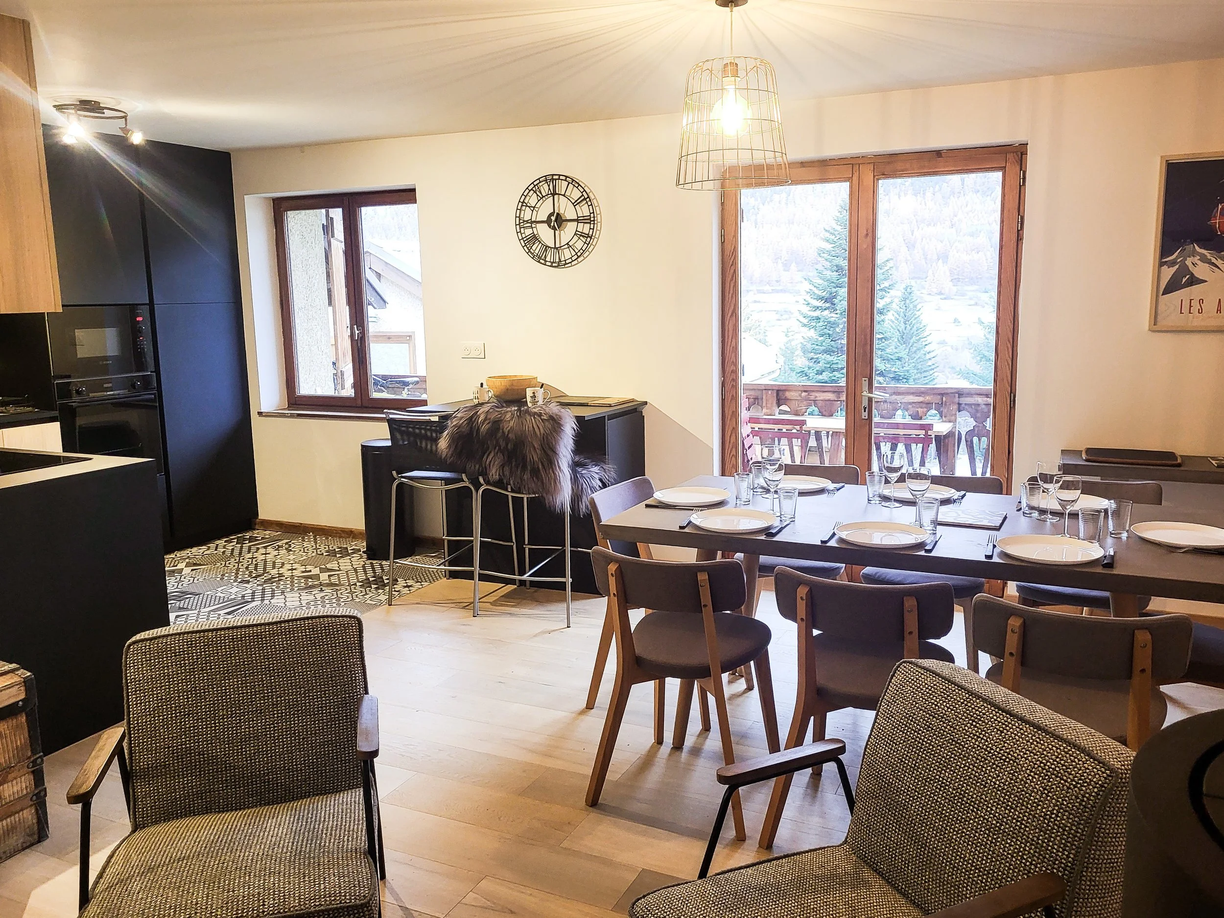 Dining area with table set for six, plates, glasses, and cutlery, chairs around the table, large windows and sliding glass door showing snowy mountain scenery, and a balcony outside. Some chairs with patterned upholstery and wooden frames, a clock on the wall, and a fur-covered stool near the breakfast bar.