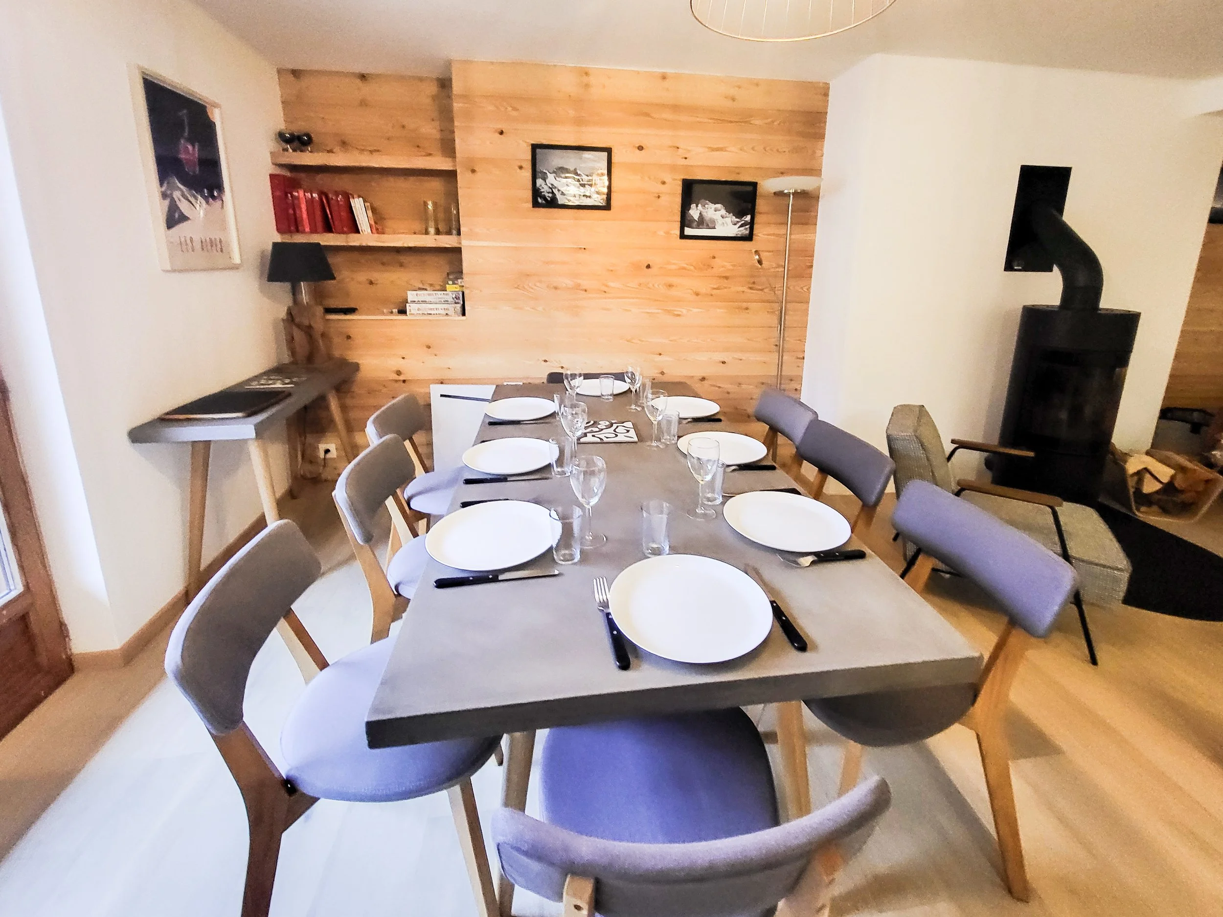 Dining room with a long table set with white plates, wine glasses, and black cutlery, surrounded by gray or beige chairs, a wood-paneled wall with framed pictures, a small bookshelf, and a wood-burning stove in the corner.